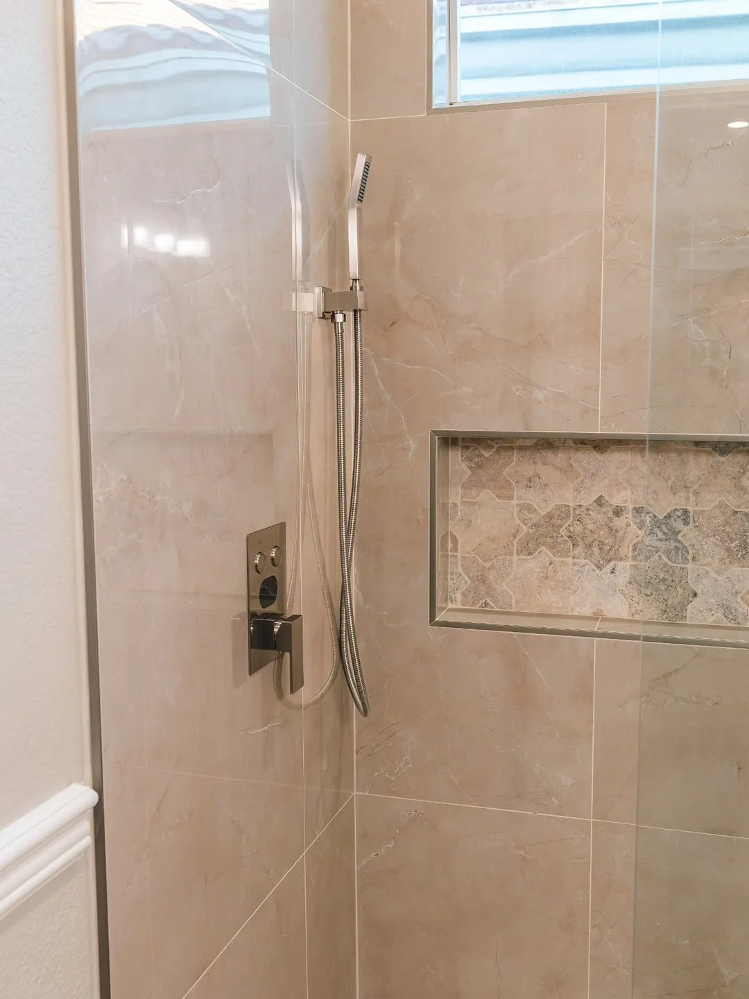 A modern shower with beige marble tiles, a built-in niche with decorative stone tiles, a handheld showerhead, and a window at the top for natural light.