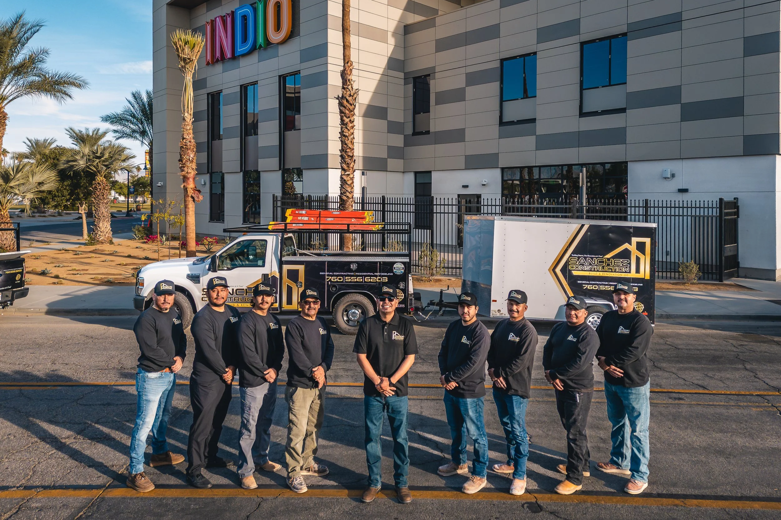 Group of nine construction workers standing in front of a Sanchez Construction truck on a city street, with a modern building and palm trees in the background.