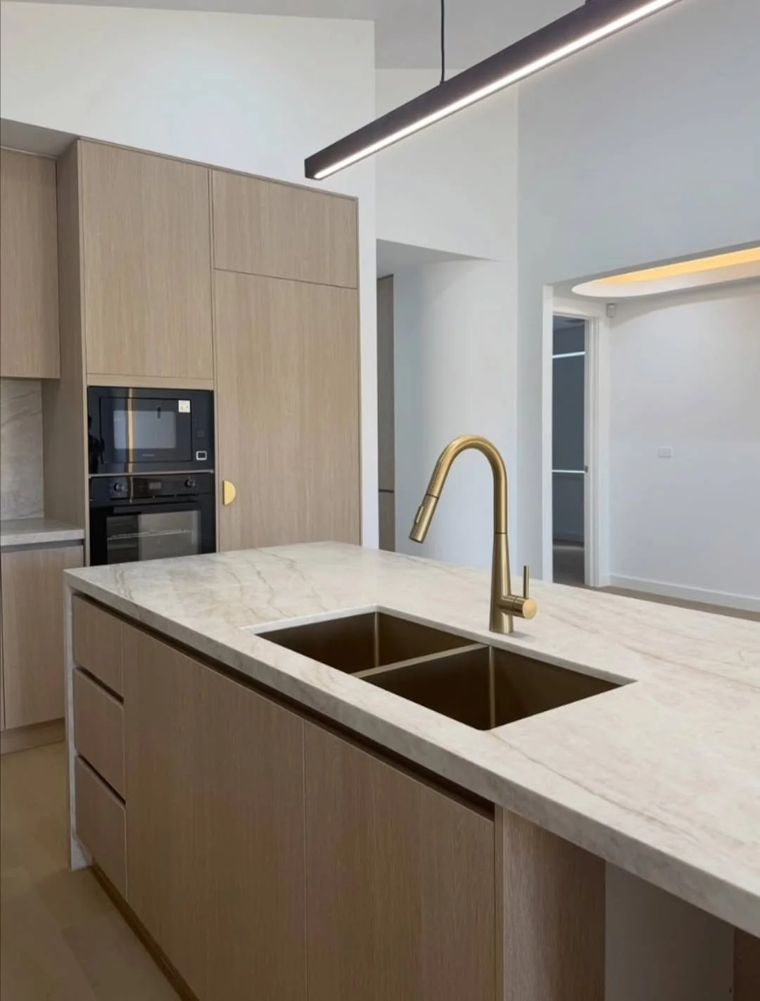 Modern kitchen with a marble island counter, gold faucet, built-in oven and microwave, light wood cabinets, and a ceiling light fixture.