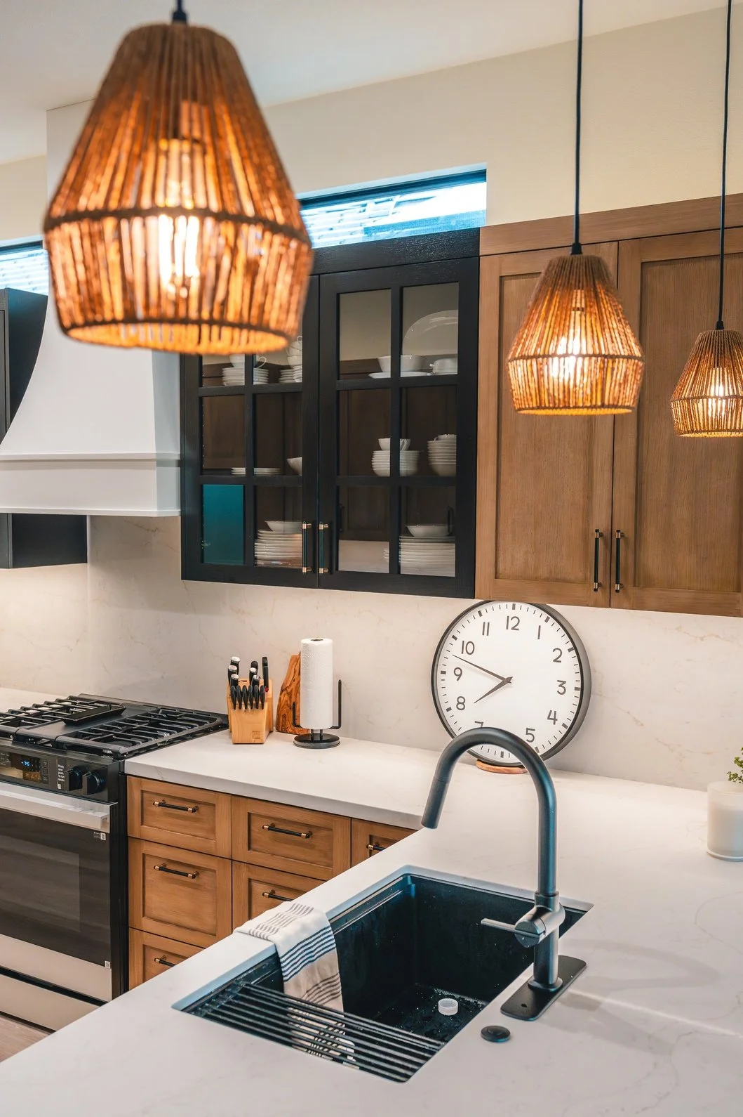 Modern kitchen with wooden cabinets, black glass door spice rack, white countertops, black sink with a striped dish towel, cypress and glass pendant lights, wall clock, paper towel holder, and a knife block.