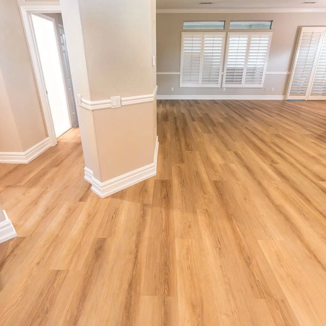 Empty living room with light wooden flooring, beige walls, white shutters on windows, and a doorway with a partly open door to another room.