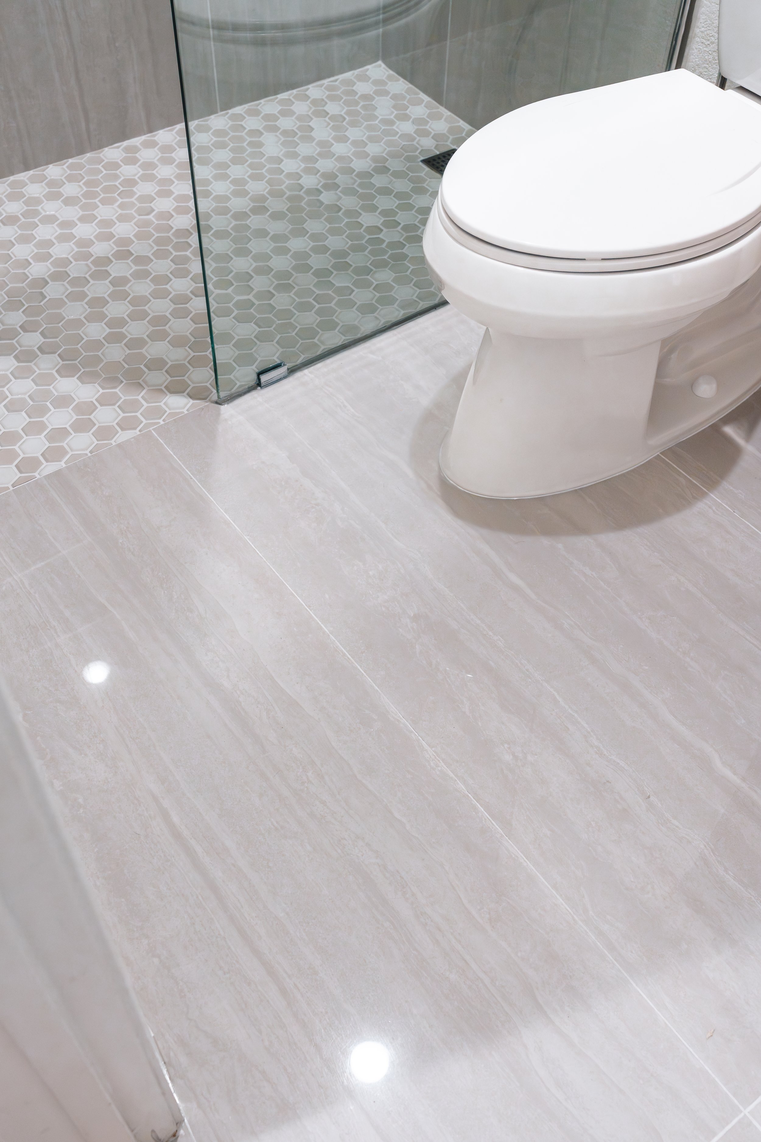 A modern bathroom with beige tile flooring, a glass-enclosed shower with hexagon tile floor, and a white toilet.