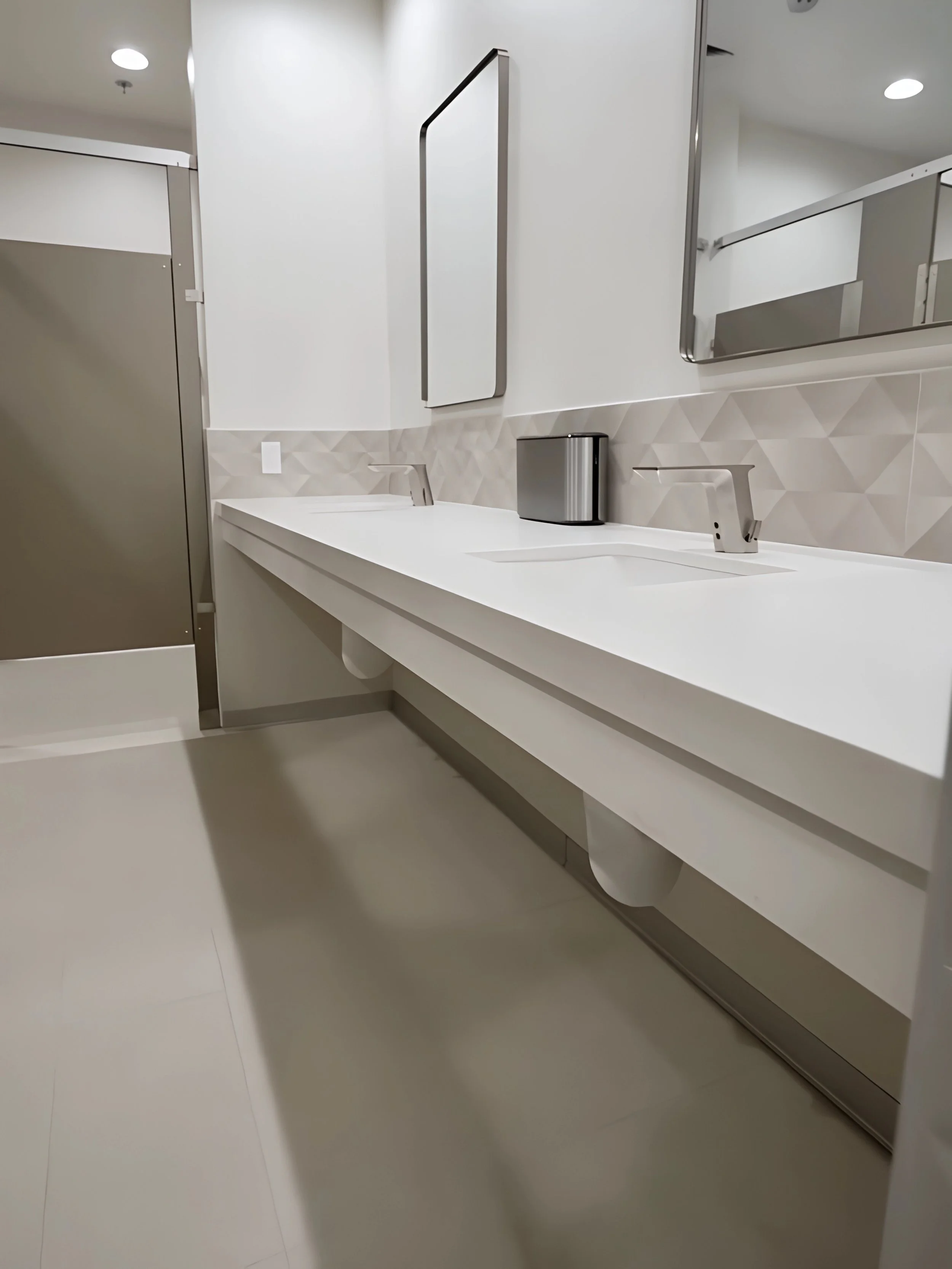 Modern white bathroom sink with two faucets, a mirror, and a trash bin, featuring a textured beige backsplash and a large rectangular mirror, with a bathroom door on the left.