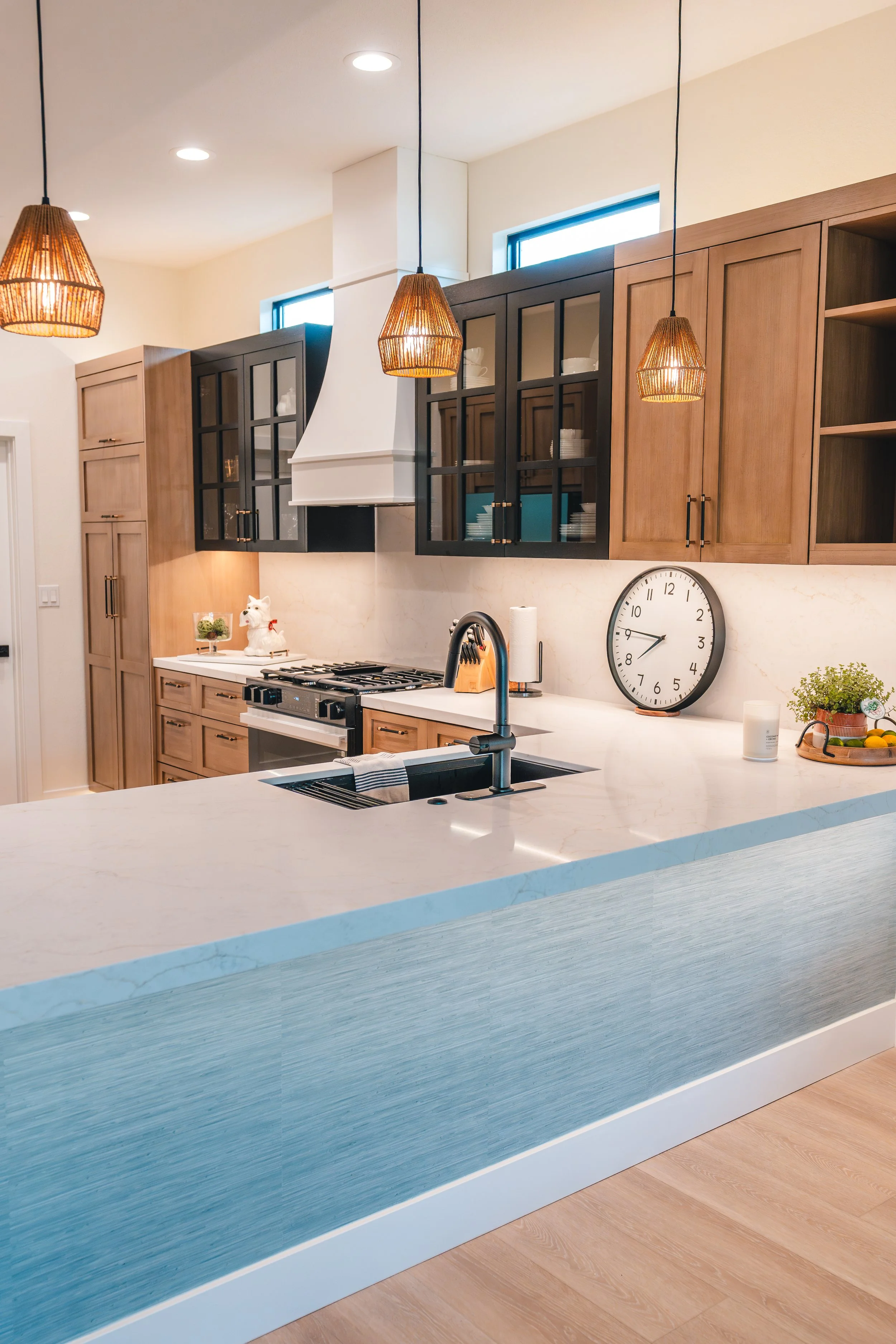 Modern kitchen with wooden cabinets, white countertops, black and glass cabinet doors, a white range hood, a clock, pendant lights, and a sink with a black faucet.