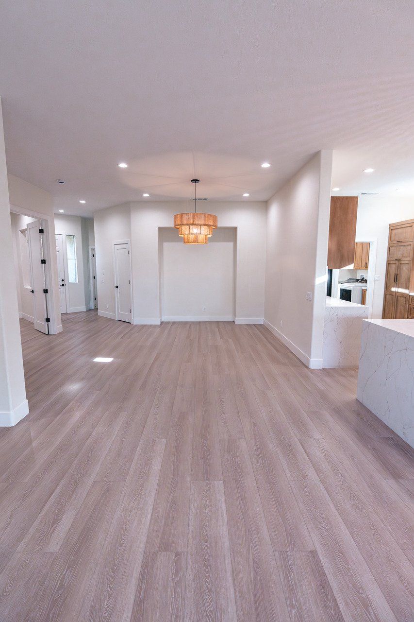 Empty living room with light wood flooring, white walls, recessed lighting, and a hanging pendant light fixture. The room opens to a kitchen area with wooden cabinets and marble countertops.