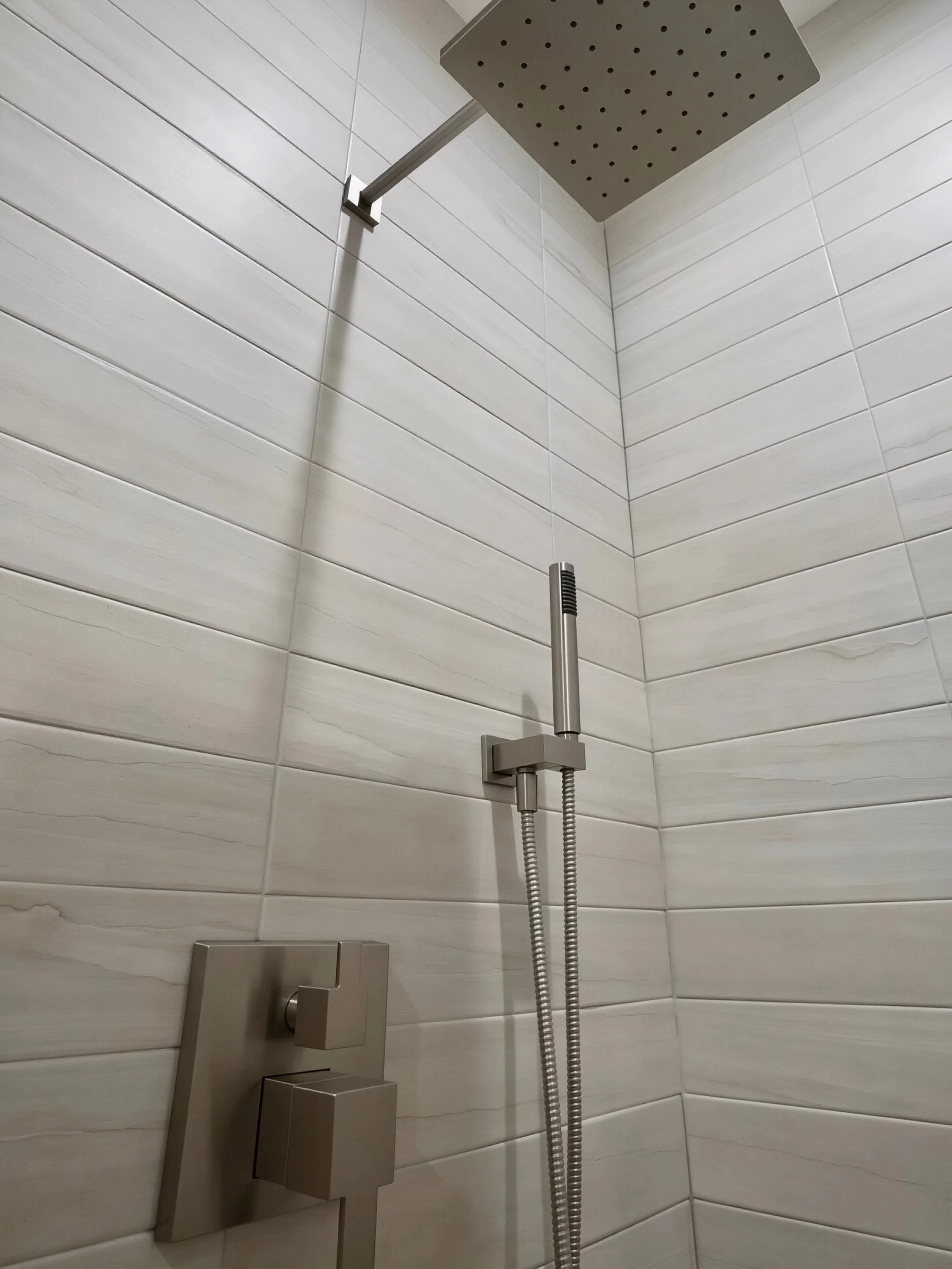 Close-up of a modern shower head and handheld shower wand with a sleek control panel, mounted on a wall with white rectangular tiles.