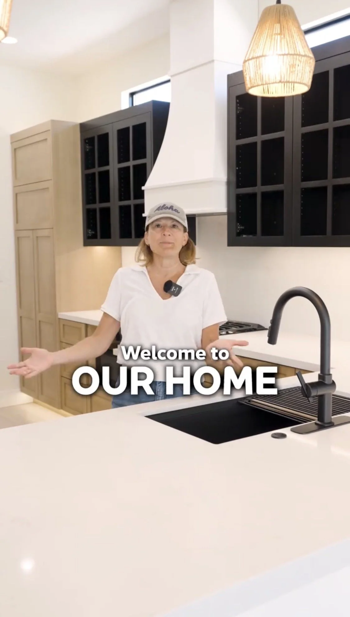 Woman in a white shirt and cap standing in a modern kitchen with black cabinets, a white countertop, and a black sink, welcoming viewers with open arms.