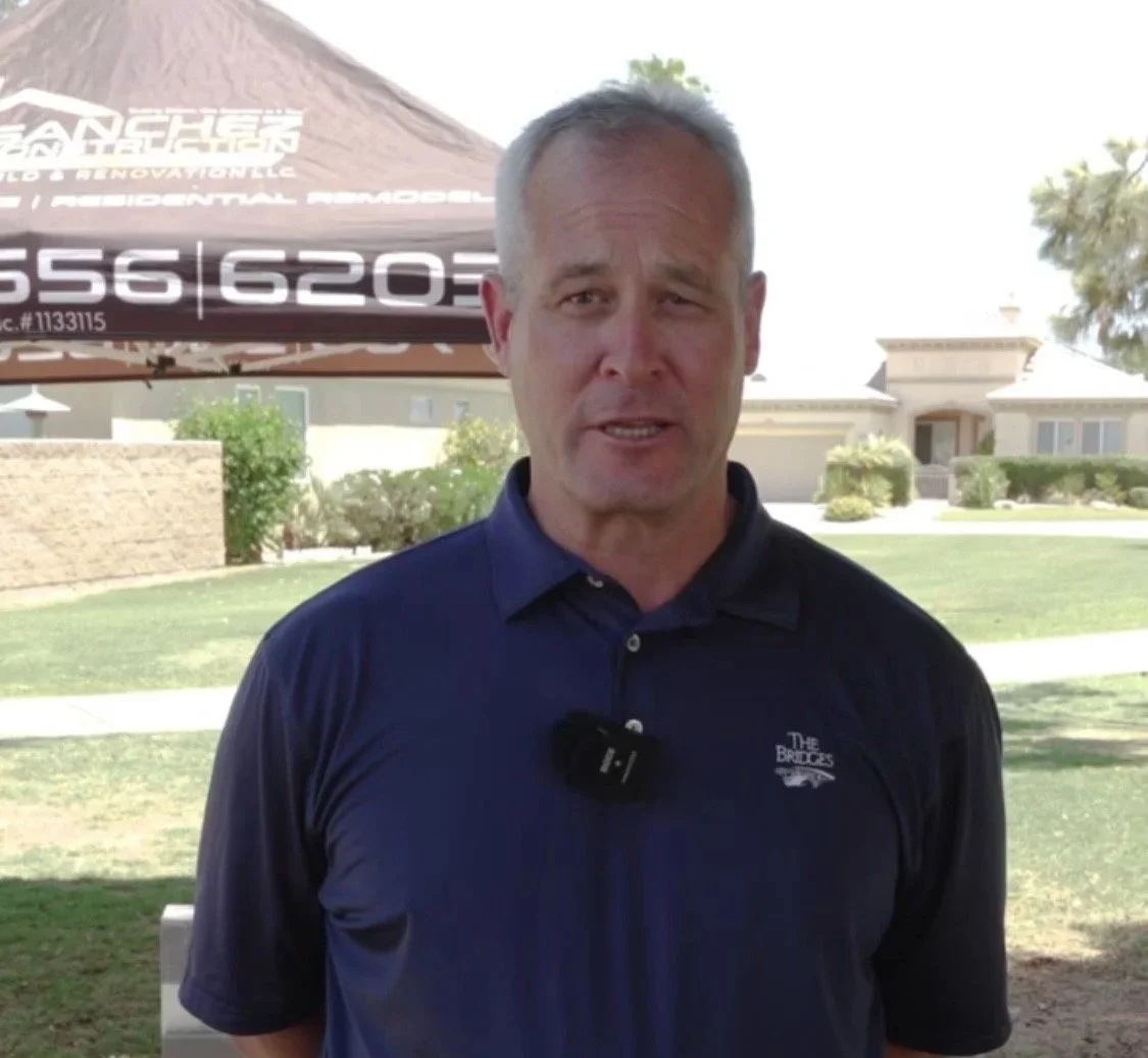 A middle-aged man standing outdoors under a canopy, wearing a navy blue polo shirt with a small logo on the chest. Behind him are houses and trees in a suburban neighborhood.