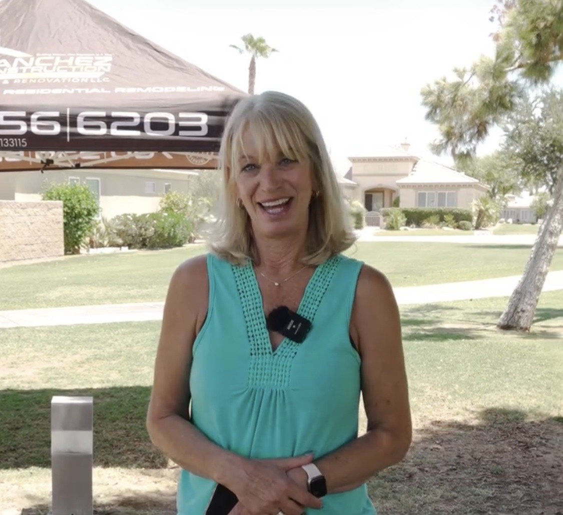 A woman with blonde hair smiling at the camera, standing outdoors under a shade with a house and trees in the background, wearing a sleeveless turquoise top and a smartwatch.