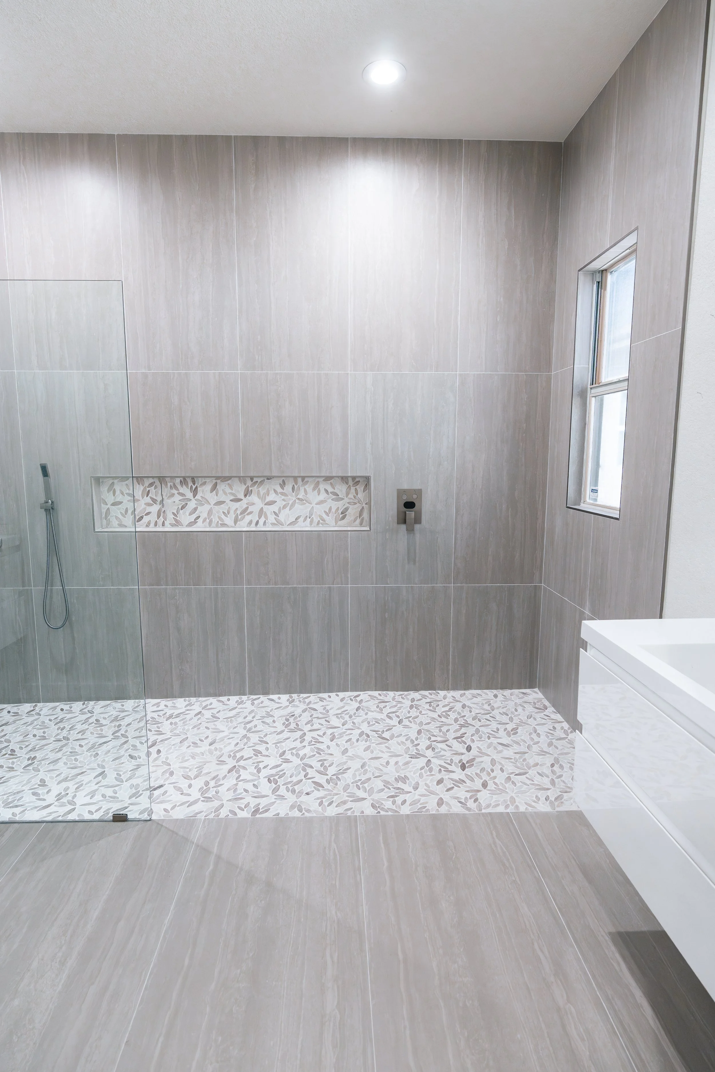 Modern bathroom with a walk-in shower, beige tiled walls, a small window, and a built-in shelf with a leaf pattern.