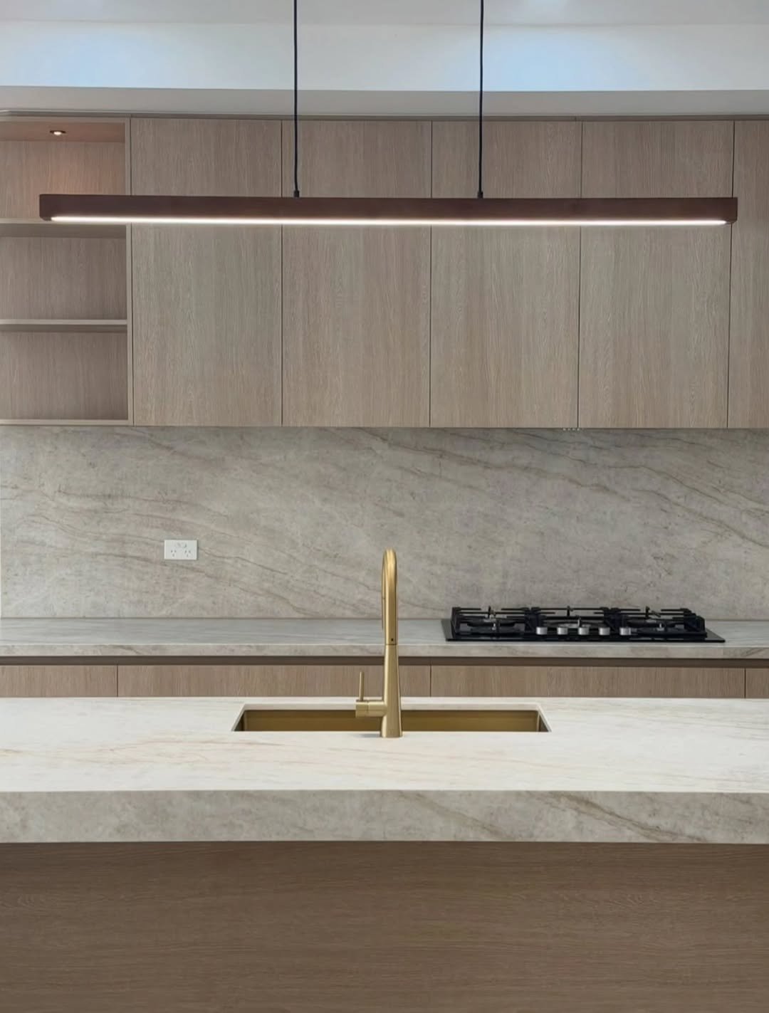 Modern kitchen with beige and wooden cabinets, a stone backsplash, a built-in stovetop, and a gold faucet over the sink in the island.