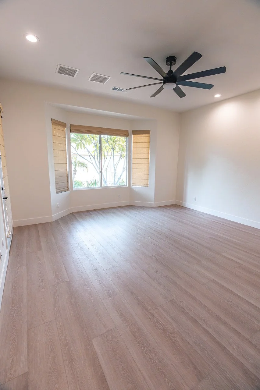 Empty room with large bay window, light wood flooring, ceiling fan, and beige window blinds.