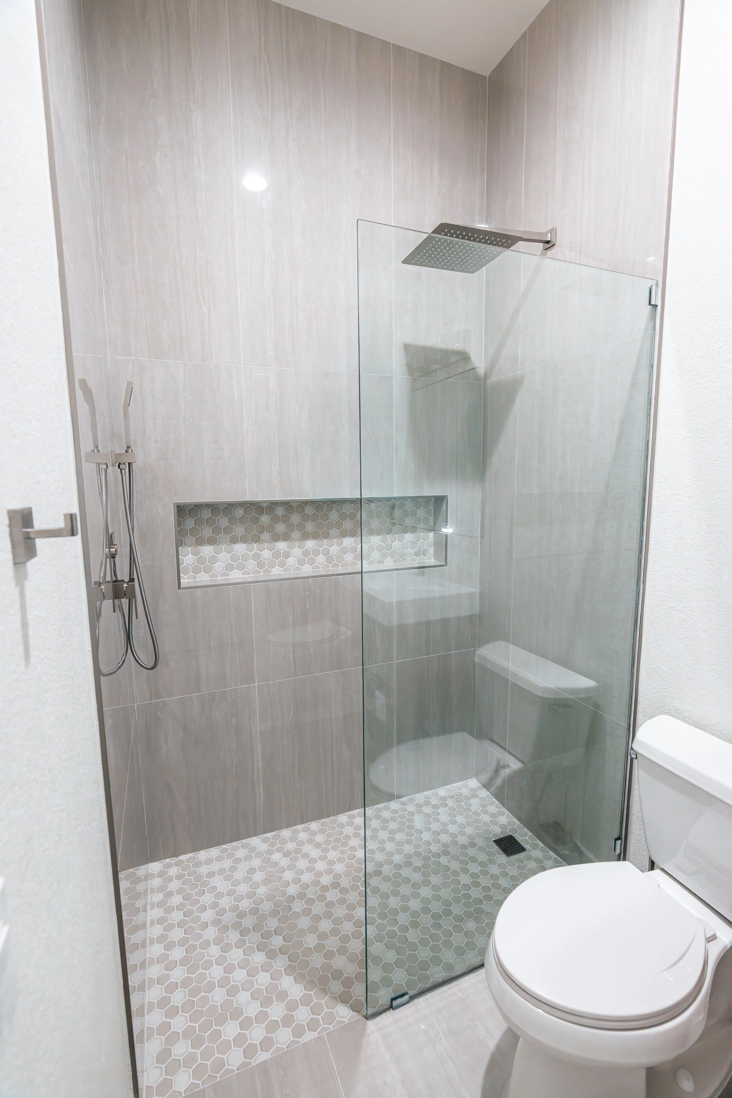 Modern bathroom with a walk-in shower enclosed by a glass partition, a rainfall showerhead, a handheld shower wand, and a built-in niche with hexagonal tiles. Toilet is visible in the foreground.