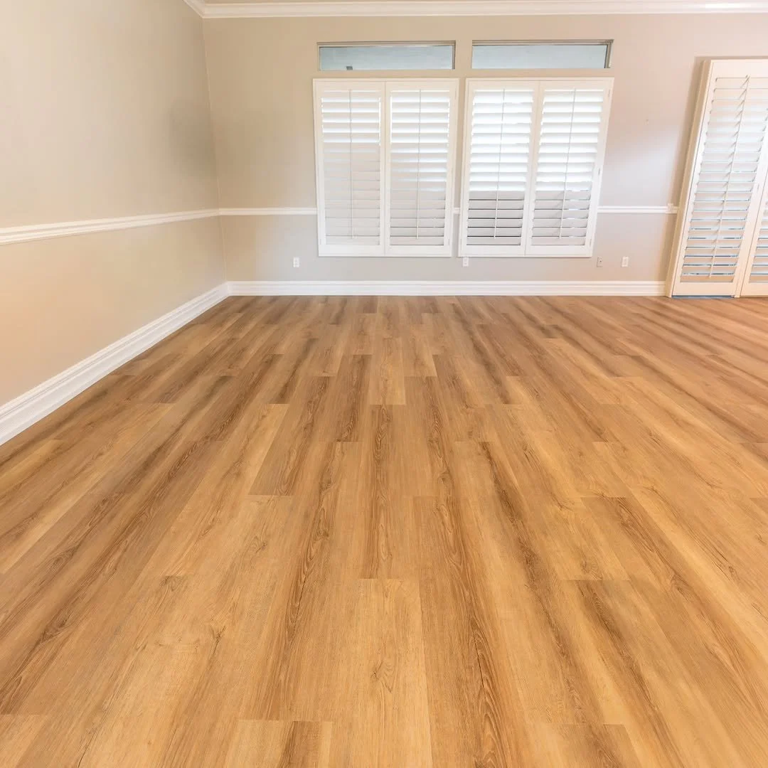 Empty room with light hardwood floors, beige walls, white trim, and large windows with white shutters.