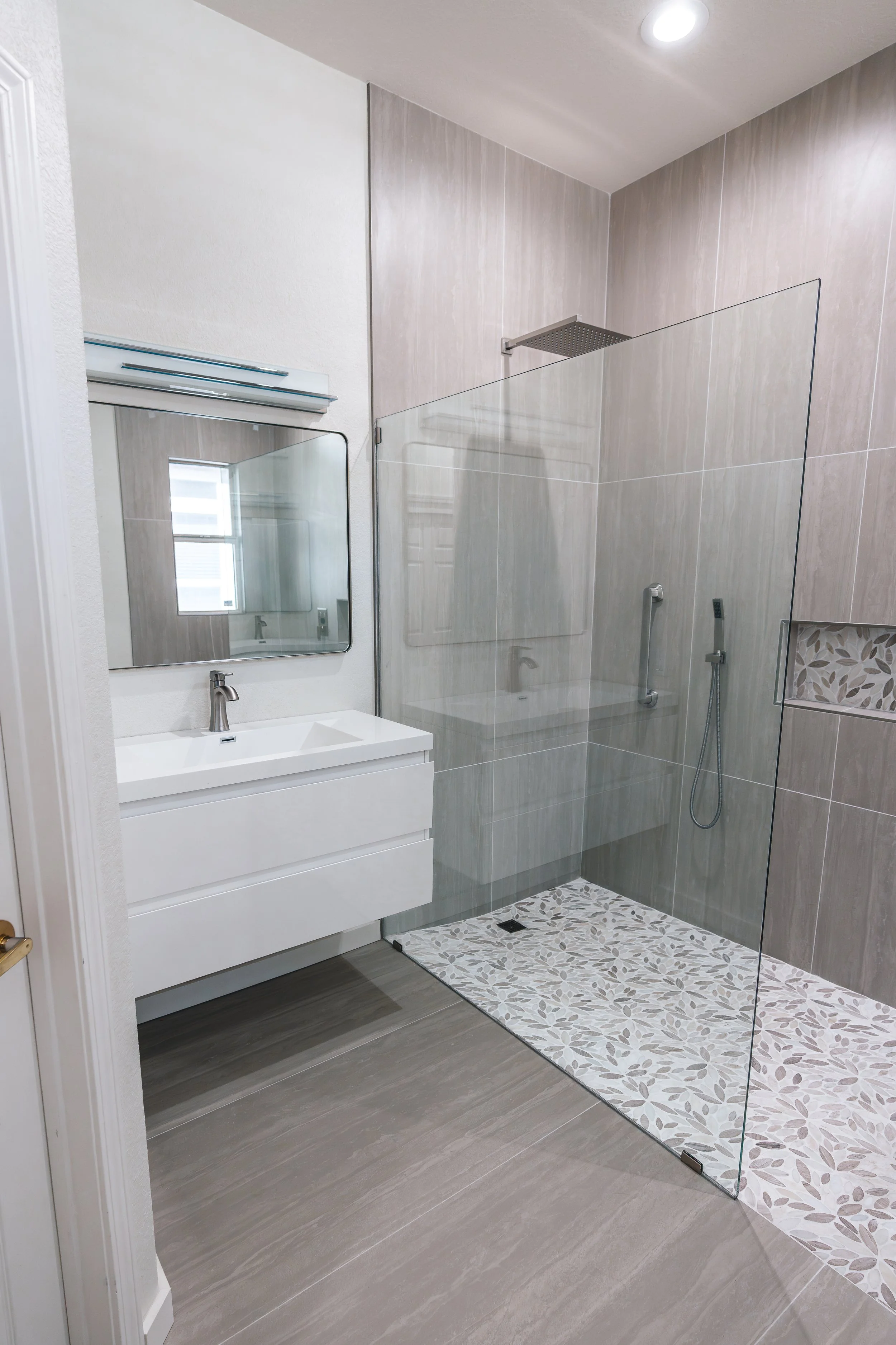 Modern bathroom with walk-in shower, glass partition, white vanity with a faucet, large wall mirror, and neutral-colored tiles with a decorative stone pattern in the shower area.