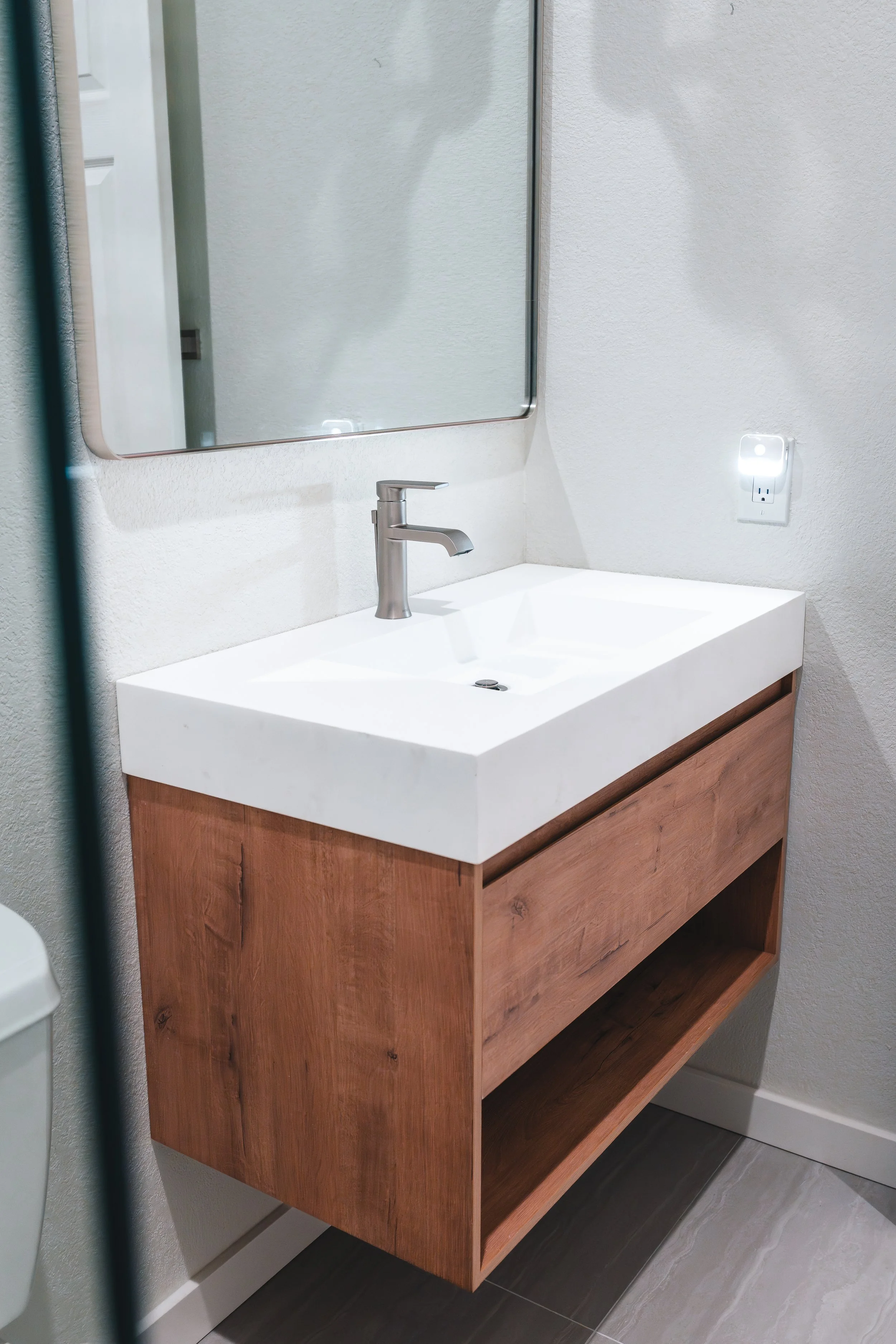 Bathroom sink with a wooden vanity, a mirror, and a wall-mounted light socket.