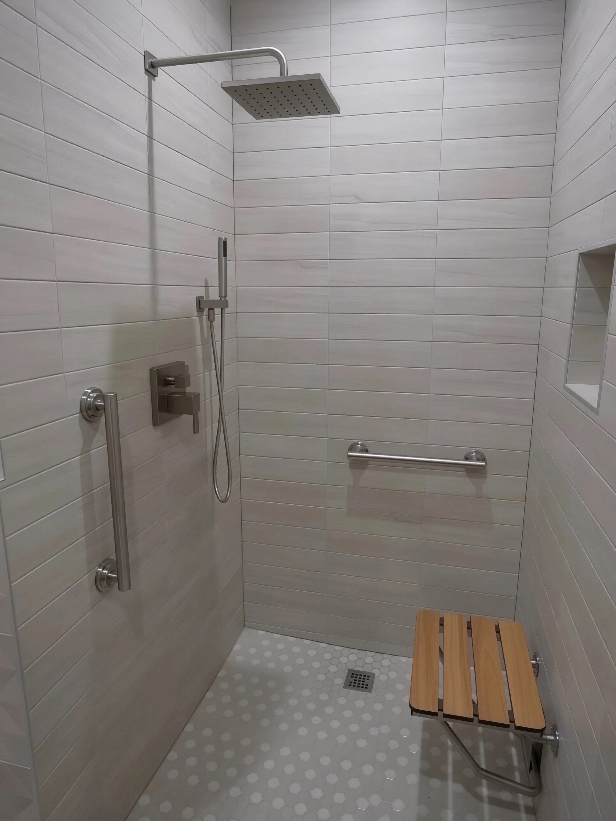 Minimalist shower stall with a large overhead showerhead, a handheld shower, two grab bars, a small built-in niche, and a wooden shower seat.