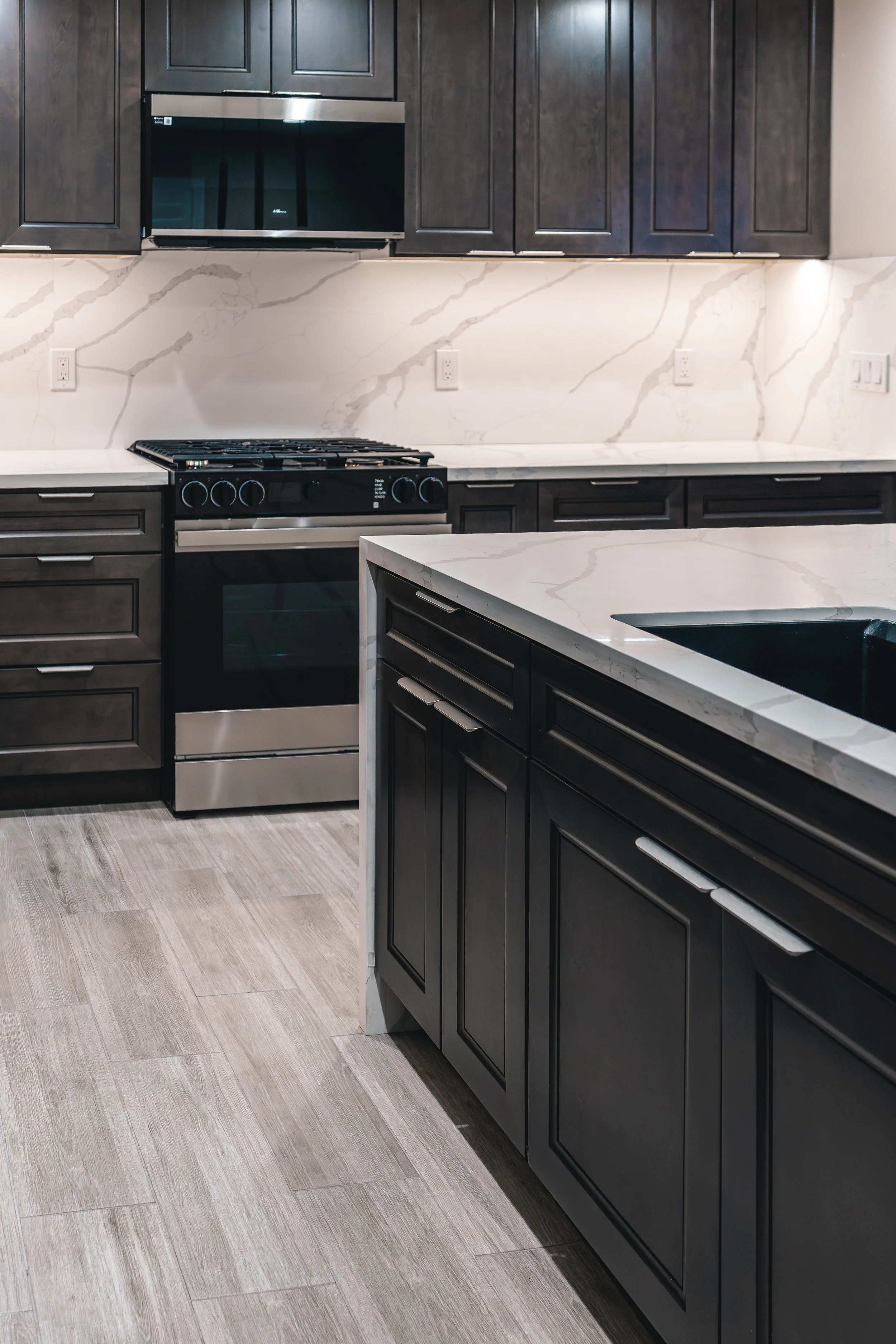 Modern kitchen with dark cabinets, marble countertops and backsplash, stainless steel oven, and tiled flooring.