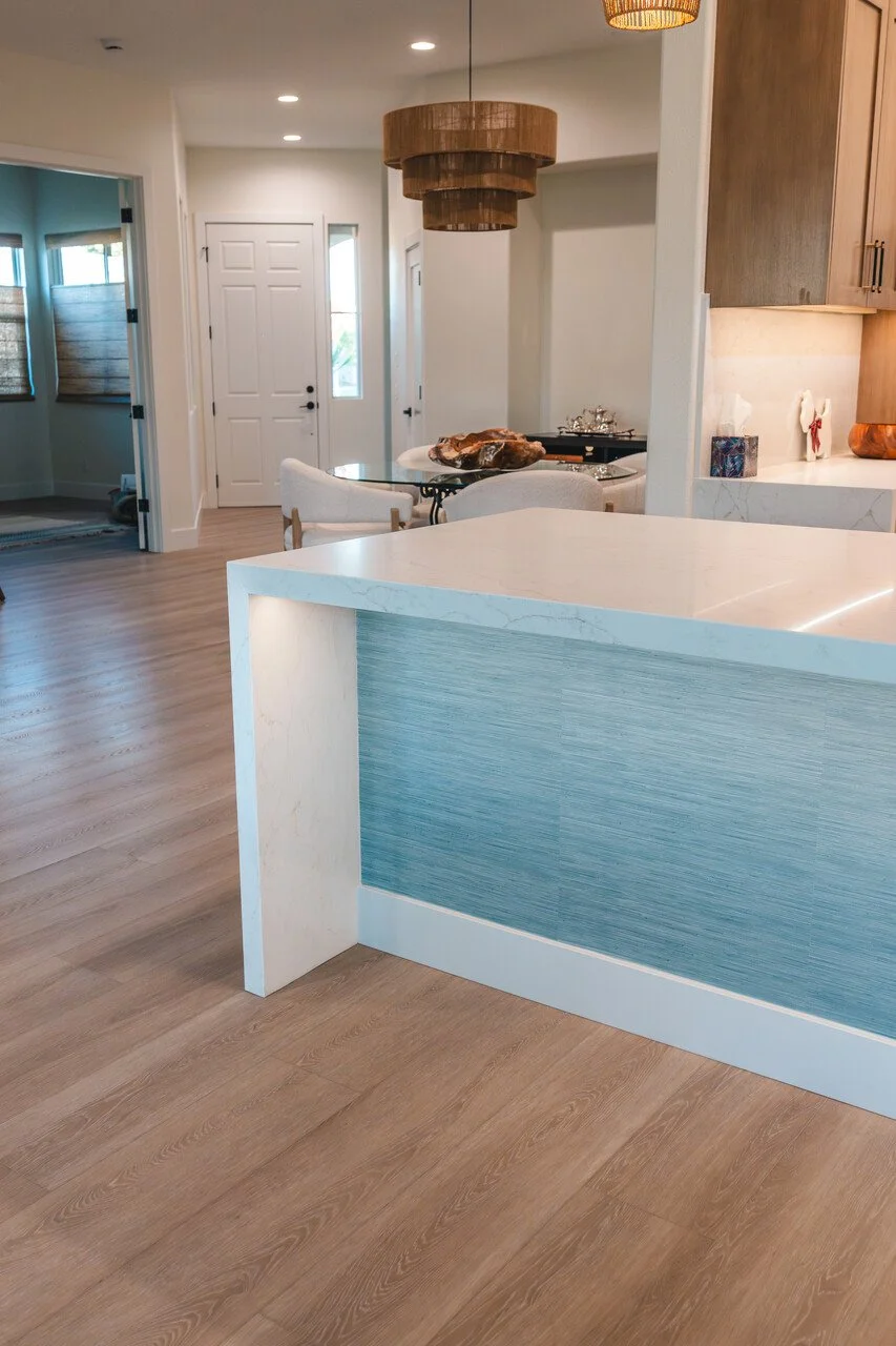 Modern open-concept kitchen and dining area with light wood flooring, white walls, and contemporary furniture. A wooden circular chandelier hangs above a dining table with white chairs. In the background, a white door and large windows with blinds ar