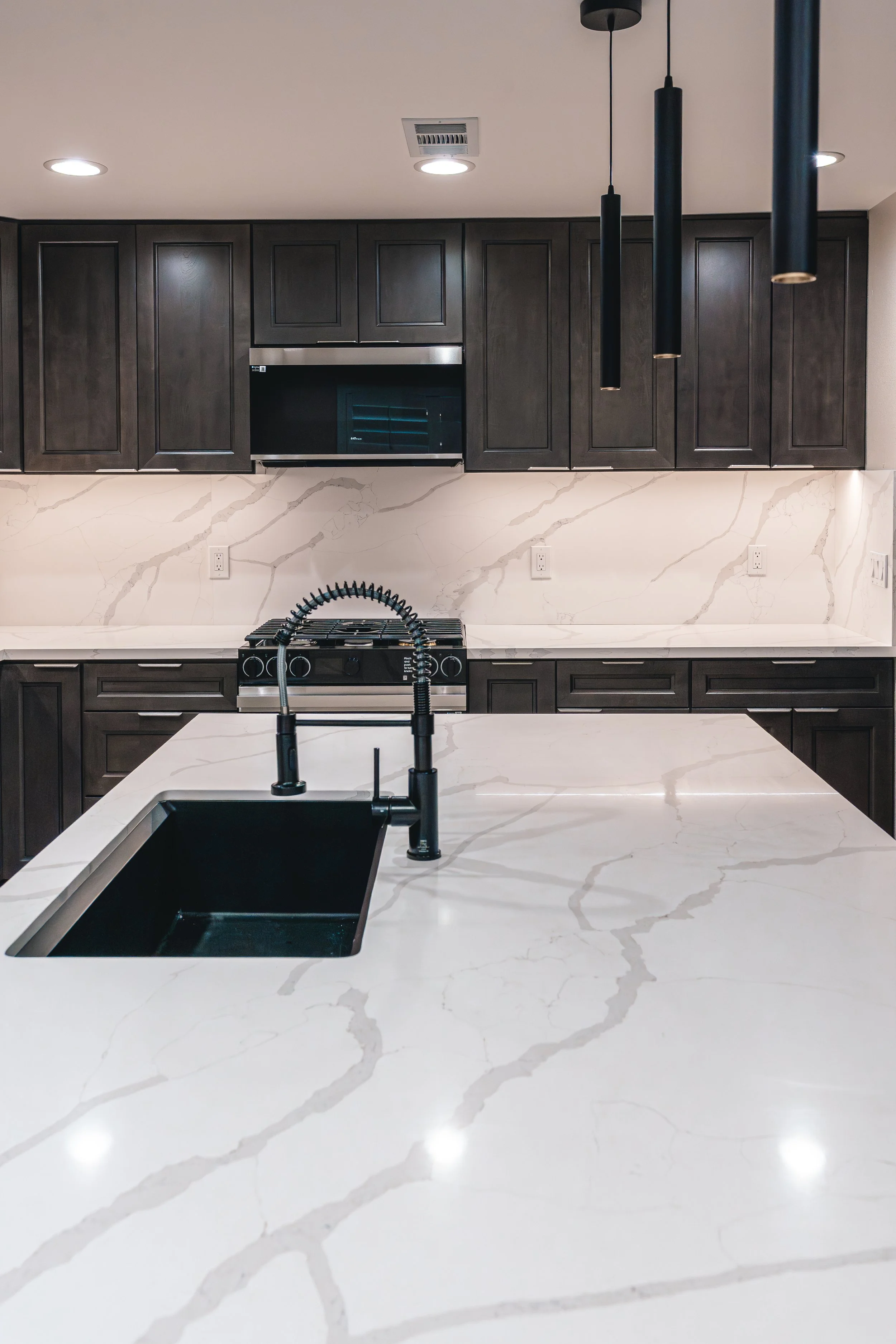 Modern kitchen with white marble countertop and backsplash, dark wooden cabinets, black faucet, and pendant lights.