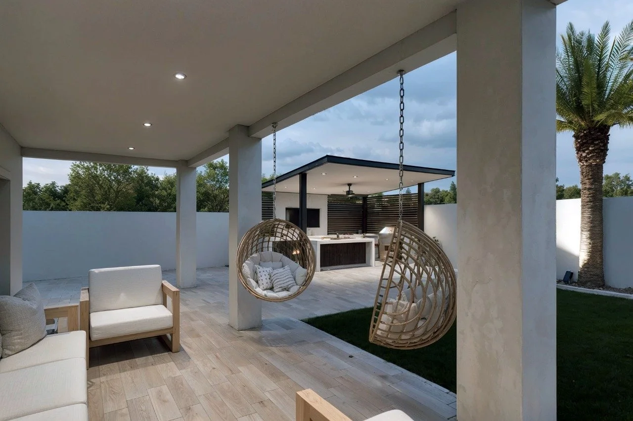 Covered outdoor patio with hanging spherical rattan chairs, outdoor sofa, and a backyard with palm tree and grassy area, featuring a modern outdoor kitchen and a white privacy fence.