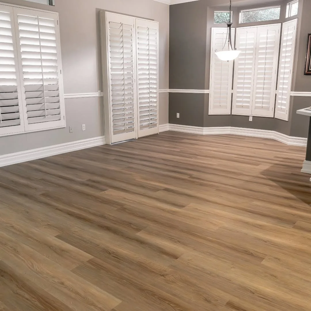 Empty living room with light wood laminate flooring, three large windows with white plantation shutters, a bay window with shutters, a hanging ceiling light, light gray walls, and white baseboards.