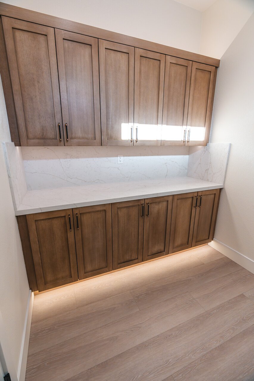 A built-in wooden cabinet with upper and lower storage, featuring vertical panel doors, a white marble countertop, and a light strip beneath the bottom cabinets illuminating the flooring.