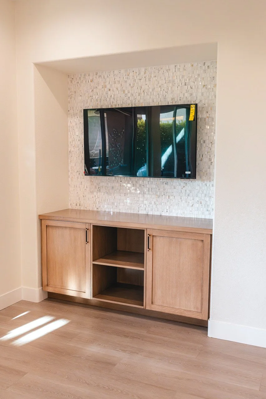 Wall-mounted flat-screen TV above a wooden cabinet with open shelves in a room with light-colored walls and wood flooring.