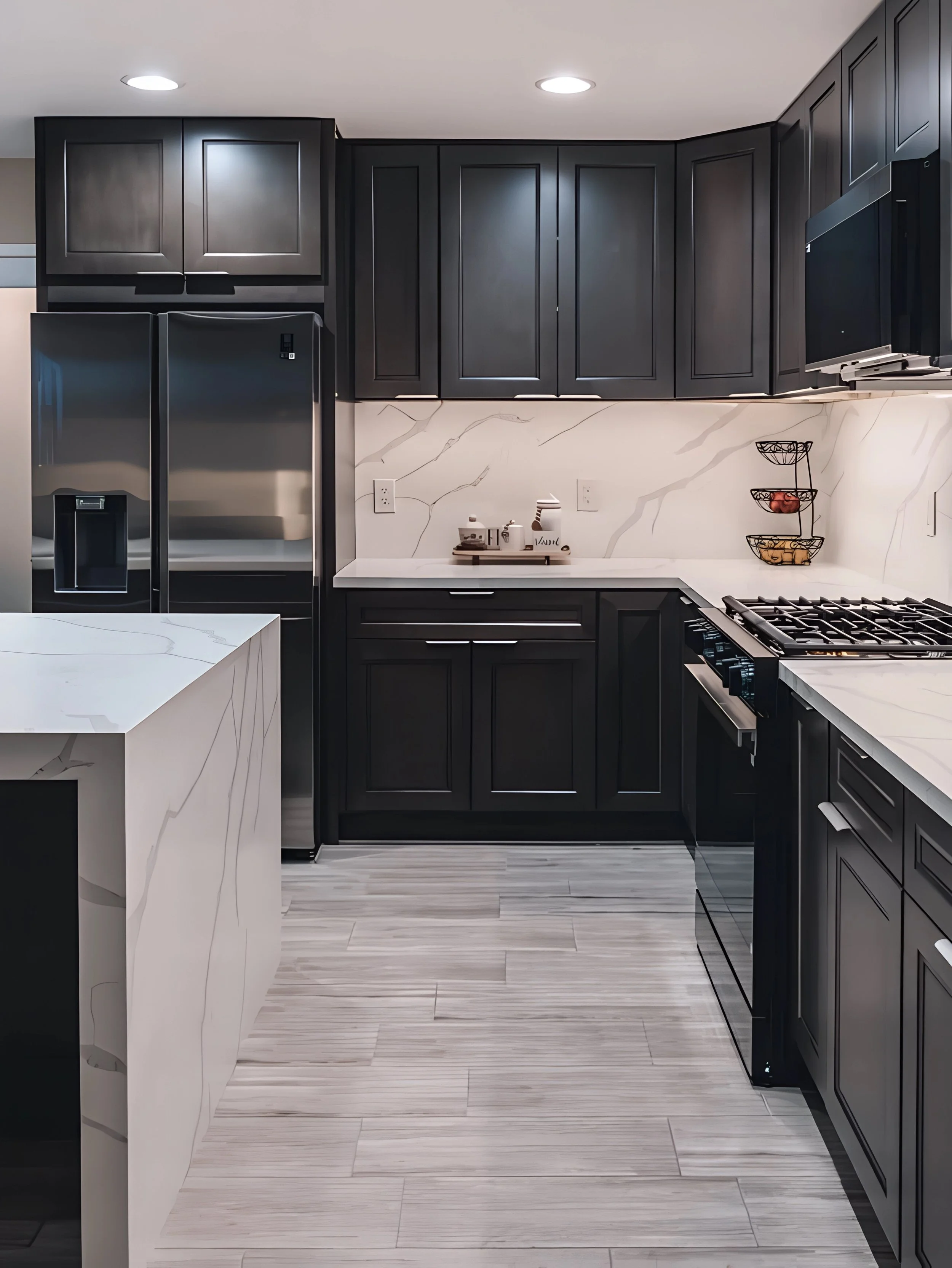 Modern kitchen with black cabinets, stainless steel refrigerator, white marble countertops, and a light wood floor.