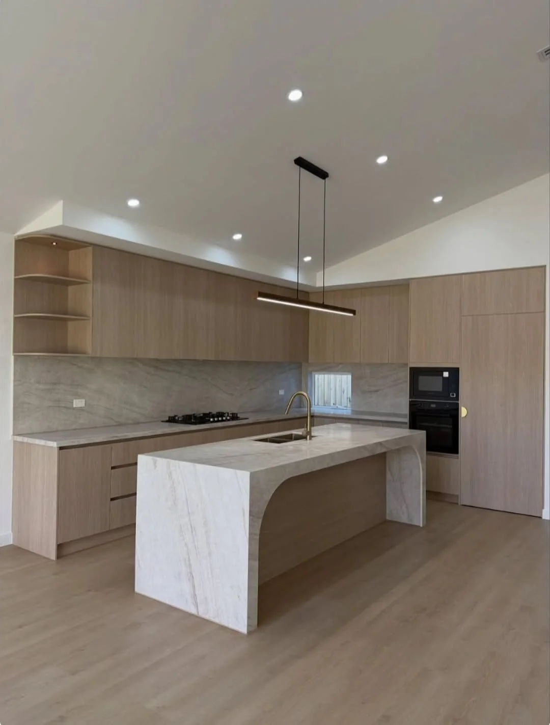 Modern kitchen with light wood cabinets, a marble island with a built-in sink, under-cabinet lighting, and a black oven and microwave unit.