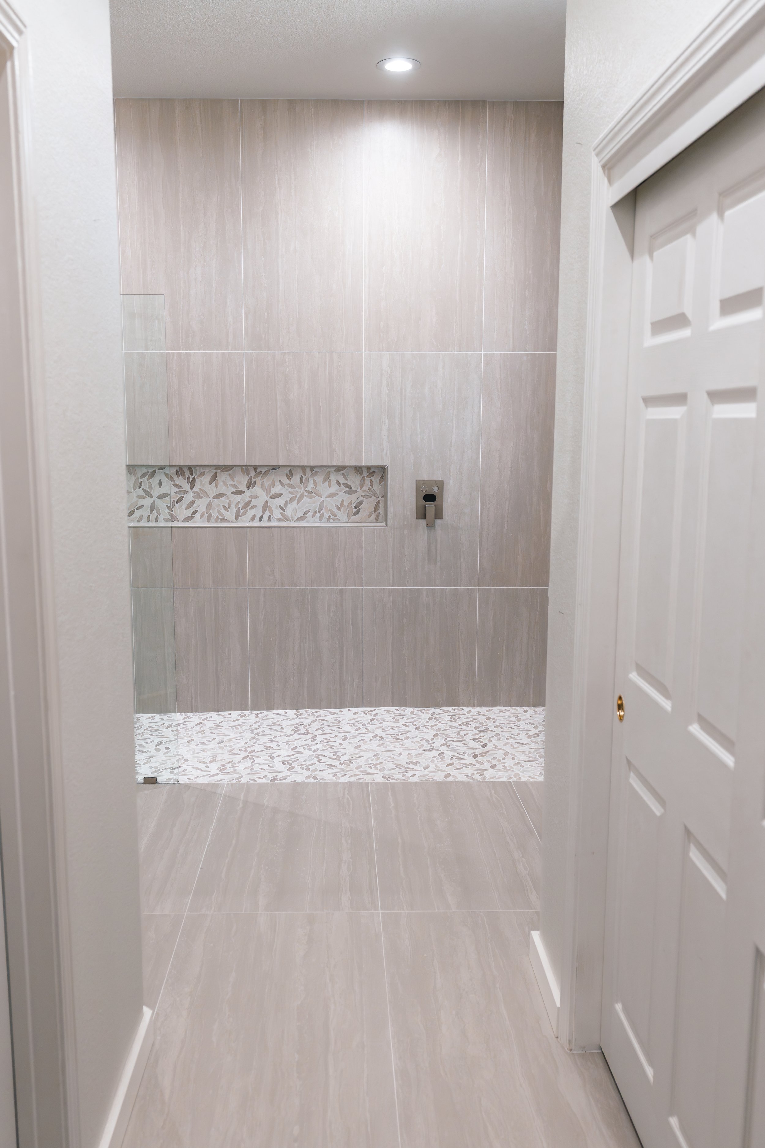 Empty shower area with beige tiles, a decorative tile strip with leaf patterns, and a small recessed shelf. White door on the right, ceiling light overhead.