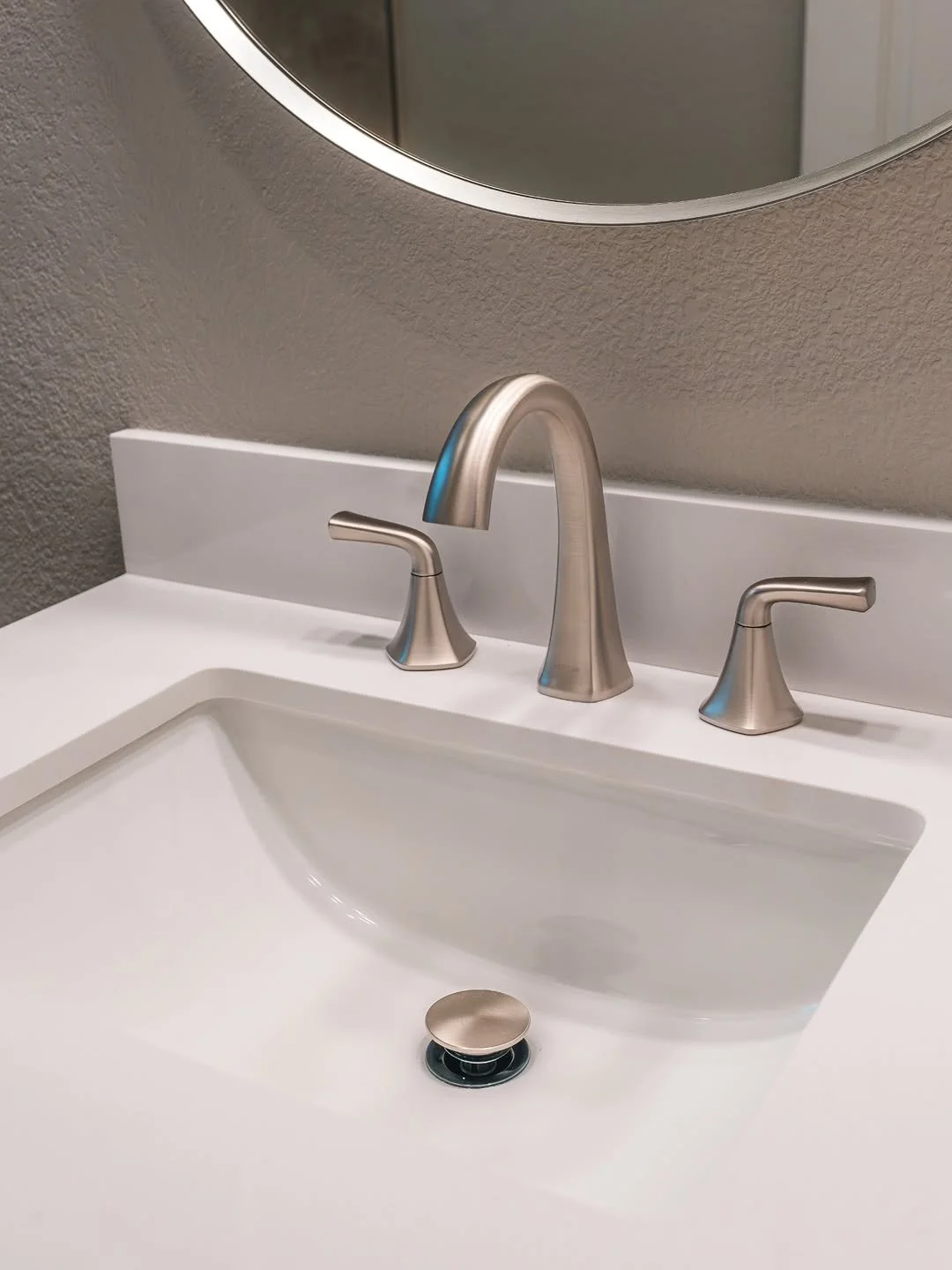 A white bathroom sink with a brushed nickel faucet and handles, a drain cover, and a mirror above. The wall behind is painted light gray.