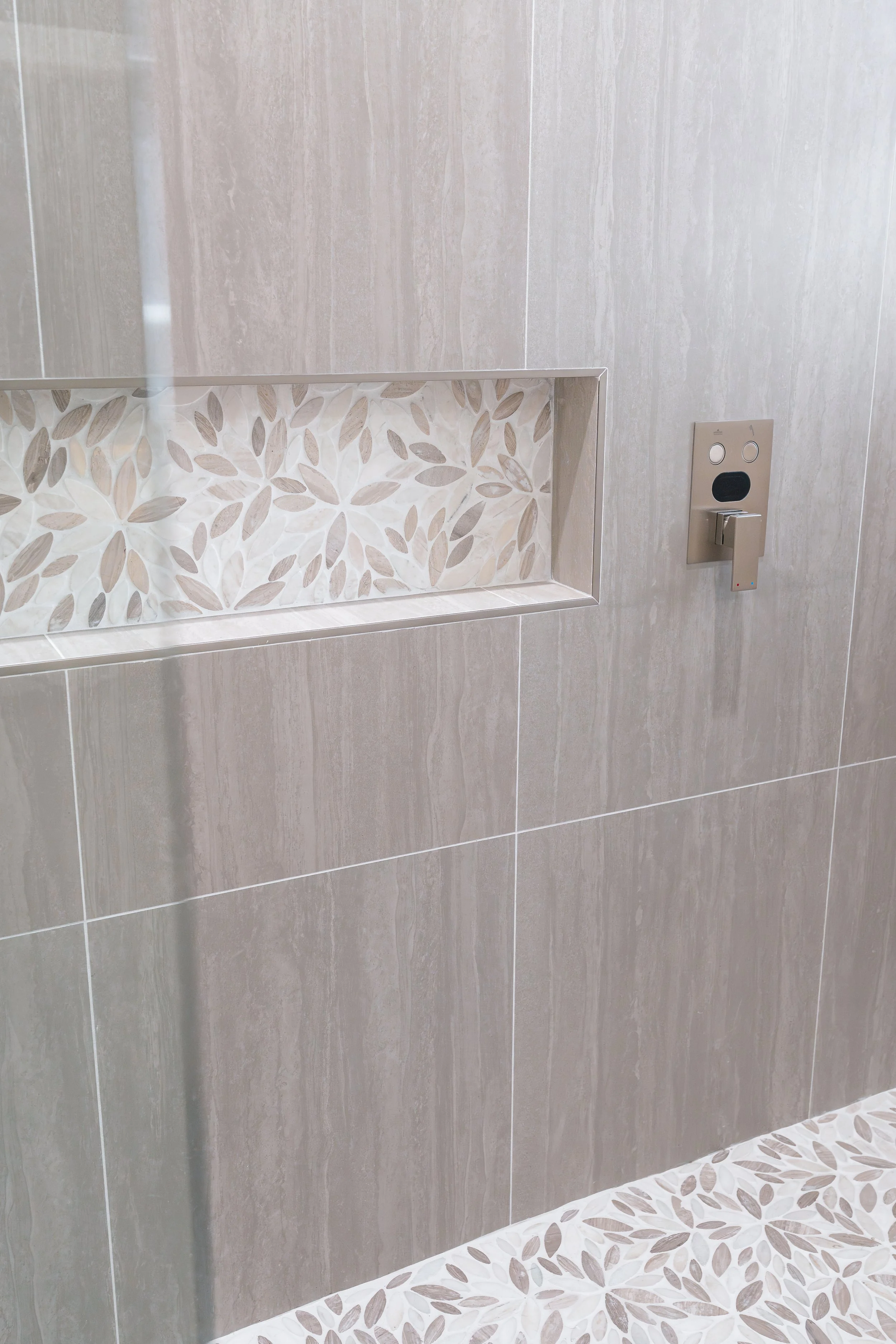 A bathroom wall with beige tiles, a decorative inset with leaf pattern, and a metallic switch or control panel.