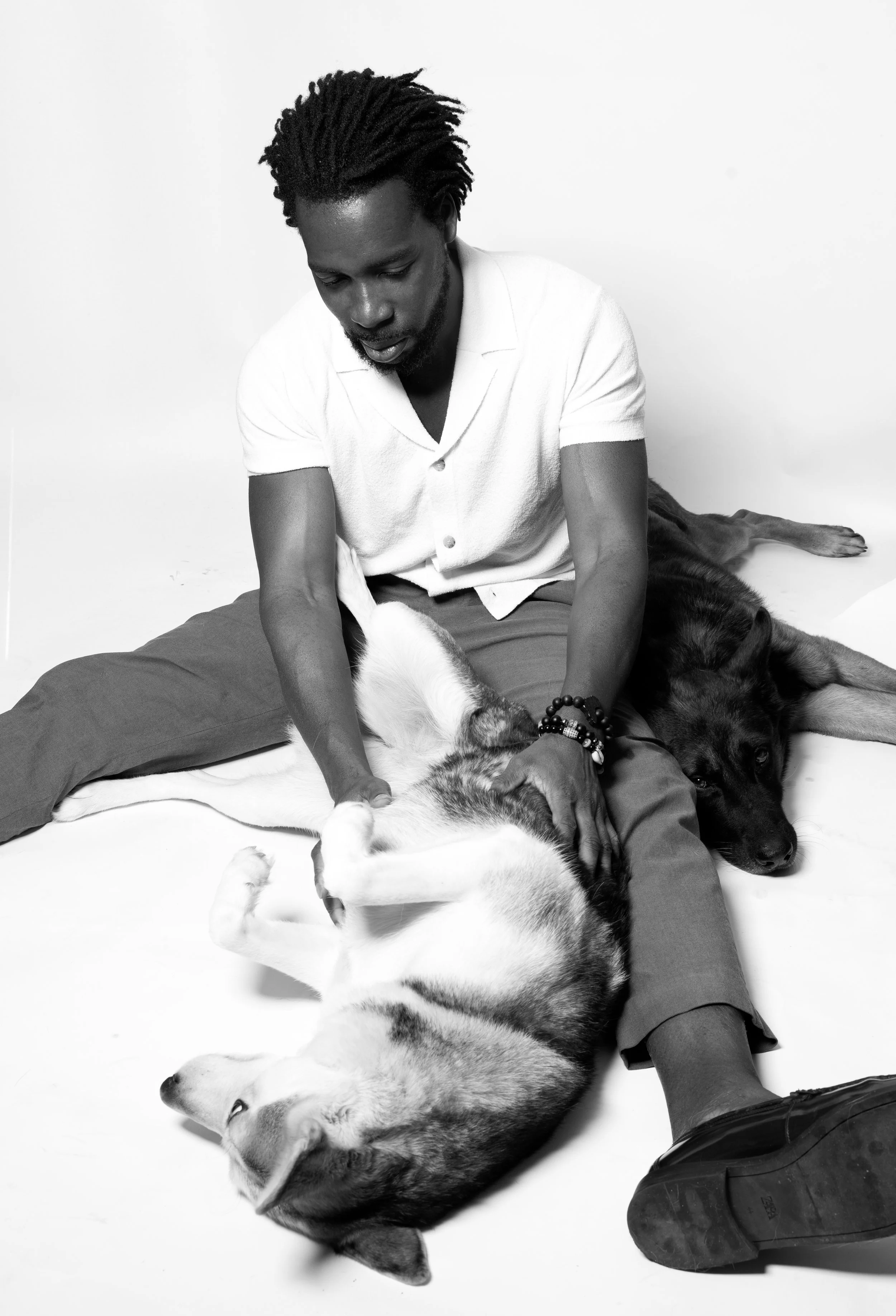 A man with dreadlocks sitting on the floor in a white shirt and pants, playing with two dogs, one lying on its back and the other lying on its side.