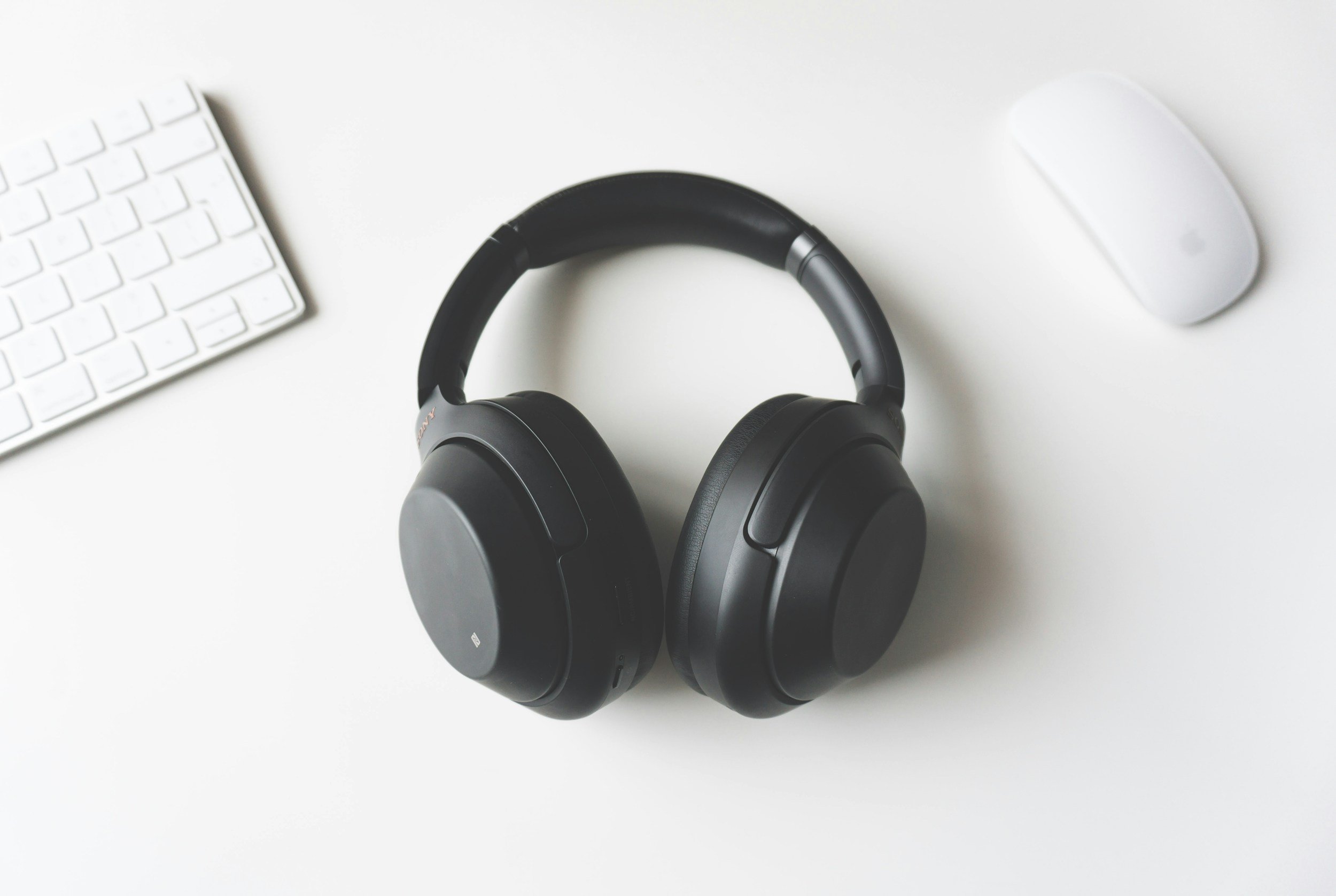 Black wireless headphones positioned in the center on a white surface with a computer keyboard partially visible on the left and a white computer mouse on the right.