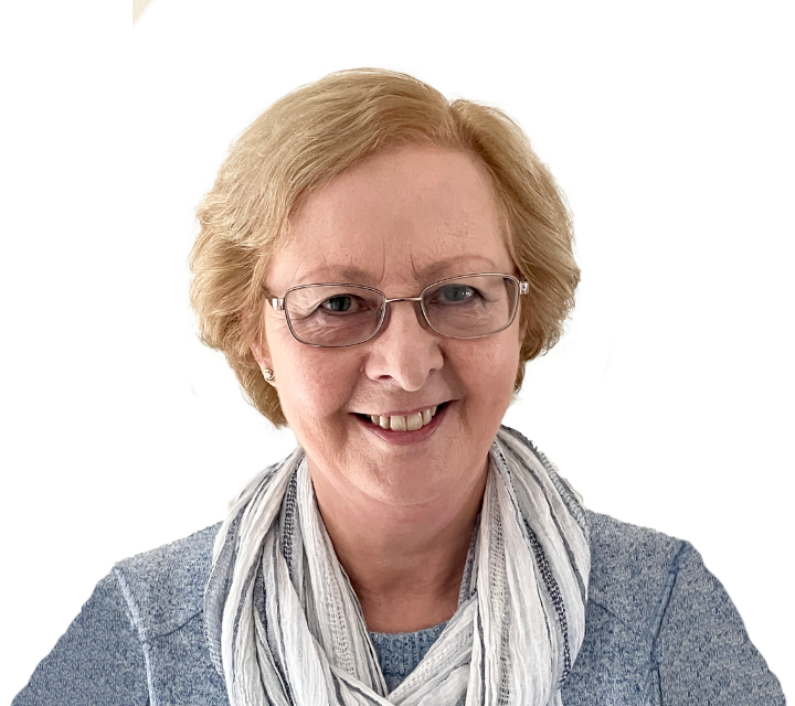 Smiling elderly woman with glasses, short blonde hair, wearing a light gray scarf and blue sweater.