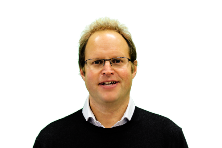 A man wearing glasses with light-colored hair, dressed in a white shirt and black sweater, smiling in front of a plain white background.