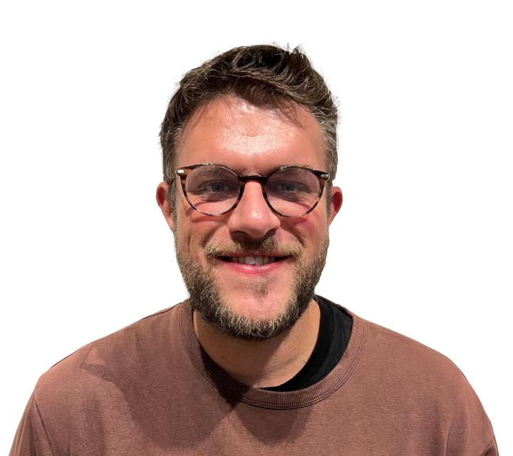 A young man with glasses and a beard smiling at the camera, wearing a brown t-shirt.