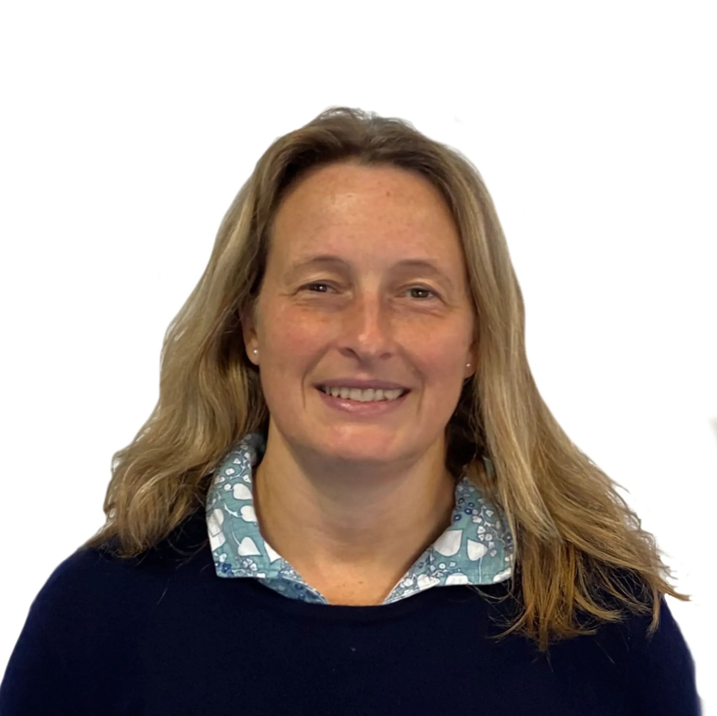 Close-up of a smiling woman with long, wavy blonde hair wearing a blue and white patterned shirt and a black sweater in front of a plain white background.