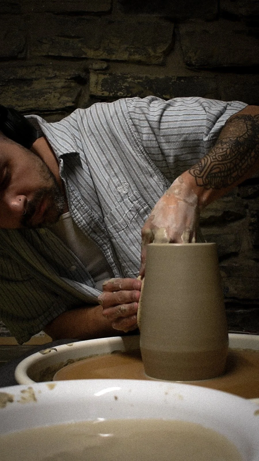 A man working on a pottery wheel, shaping a clay vase with his hands. The man has tattoos on his arm, and he is wearing a striped shirt. The background features a stone wall.
