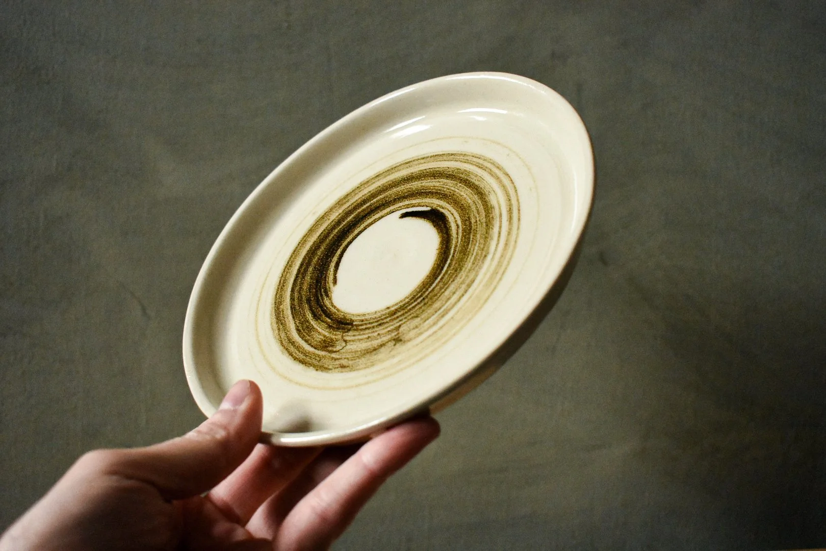 A hand holding a ceramic plate with a brown spiral pattern on a beige background.
