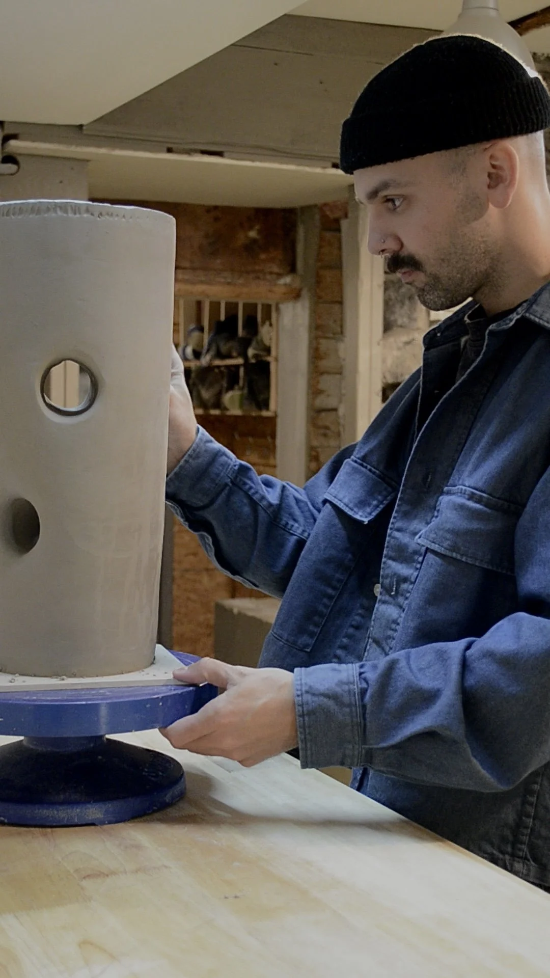 A man wearing a black beanie and denim jacket working on a pottery wheel shaping clay into a ceramic piece in a workshop.