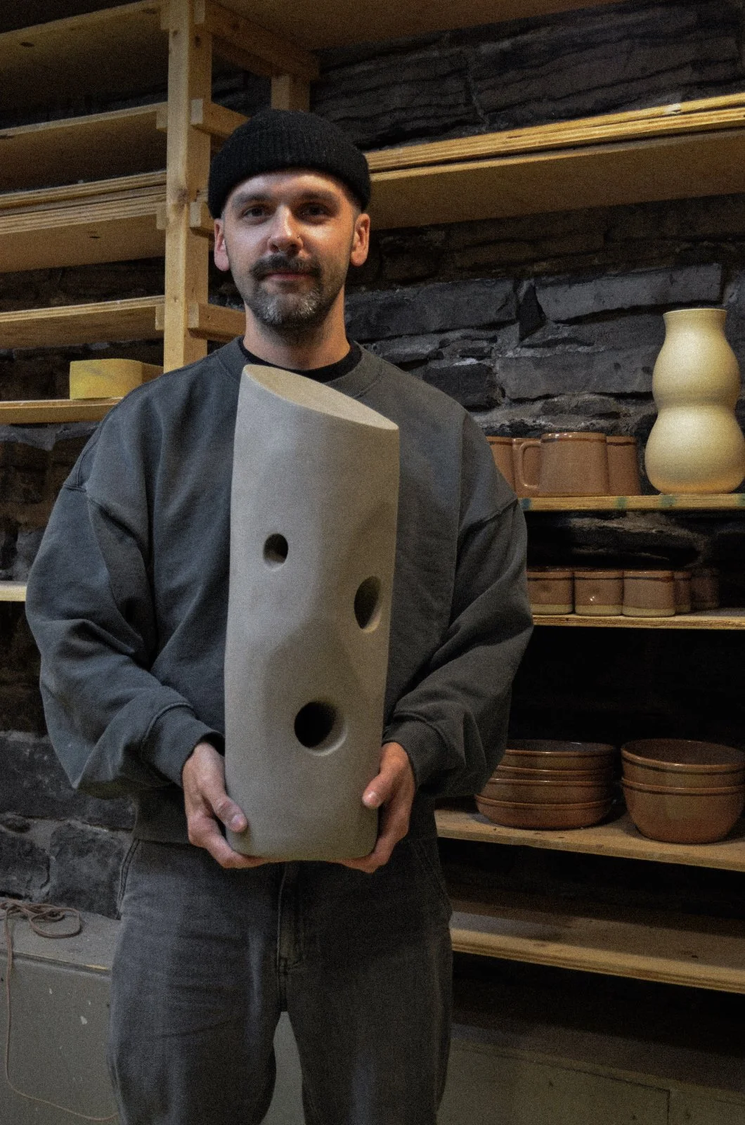 Man in grey sweatshirt and black beanie holding a ceramic cheese board with holes, standing in a pottery studio with shelves of pottery in the background.
