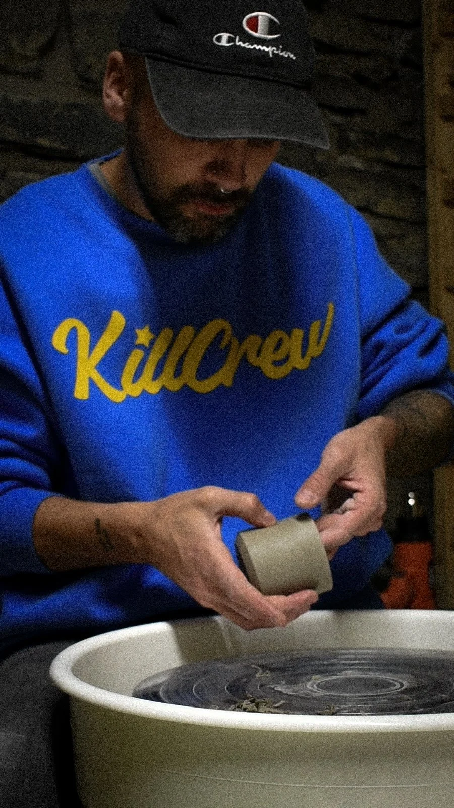 A person with tattoos on their wrists wearing a blue sweatshirt with yellow text and a black Champion cap, using a pottery wheel to shape clay.