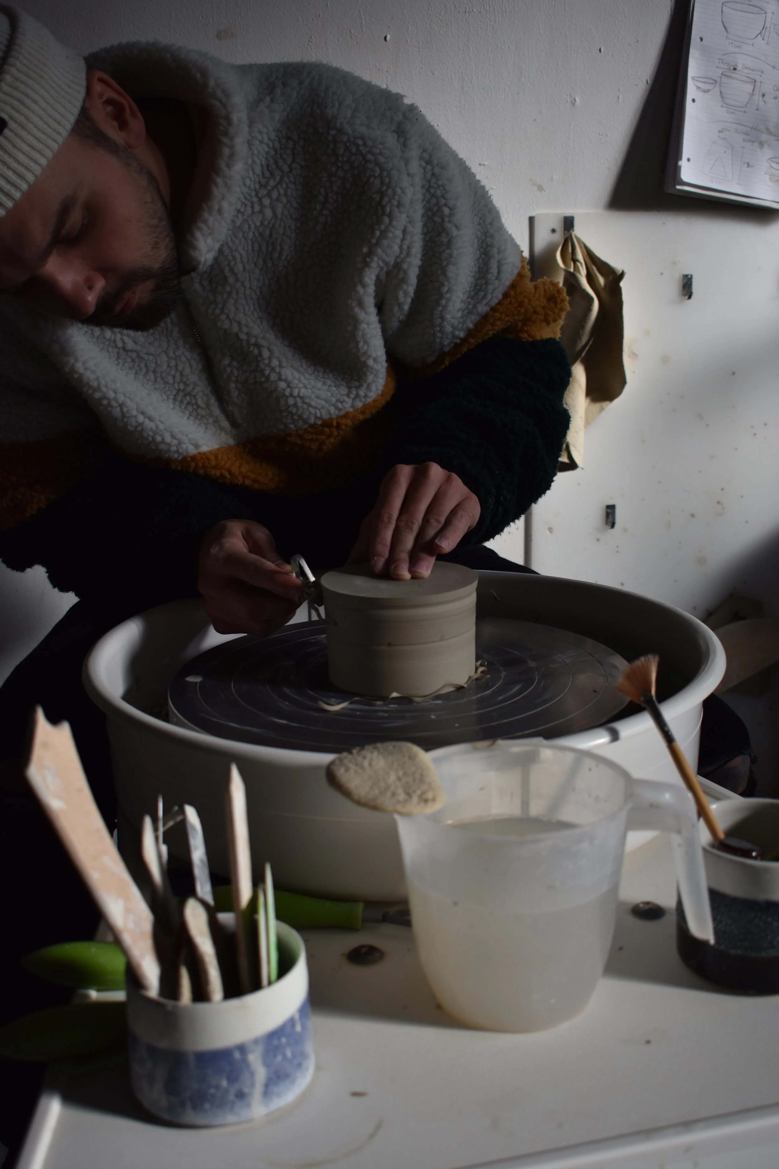 A person working on a pottery wheel, shaping clay with their hands. The person is wearing a fluffy, multicolored jacket and a beanie. Various pottery tools and supplies are on the table nearby.