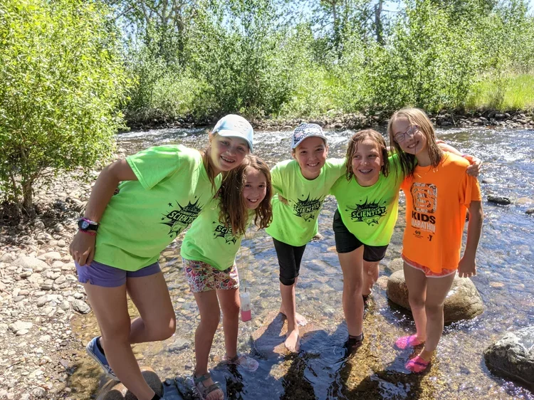 Five young girls in bright t-shirts standing in a shallow stream with rocks, surrounded by green trees on a sunny day.