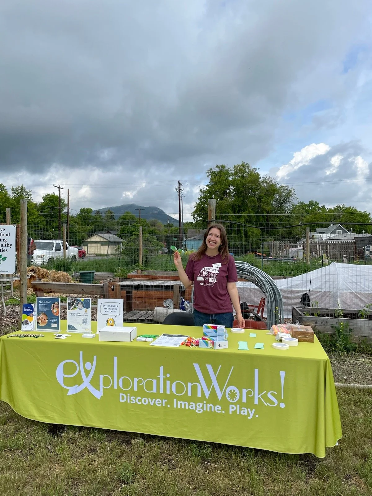 A smiling woman standing behind a bright green tablecloth at an outdoor booth for Exploration Works, which features the slogan 'Discover. Imagine. Play.' The table has various informational pamphlets, small items, and a raffle sign. The background shows a fenced garden area, trees, parked cars, and a cloudy sky.
