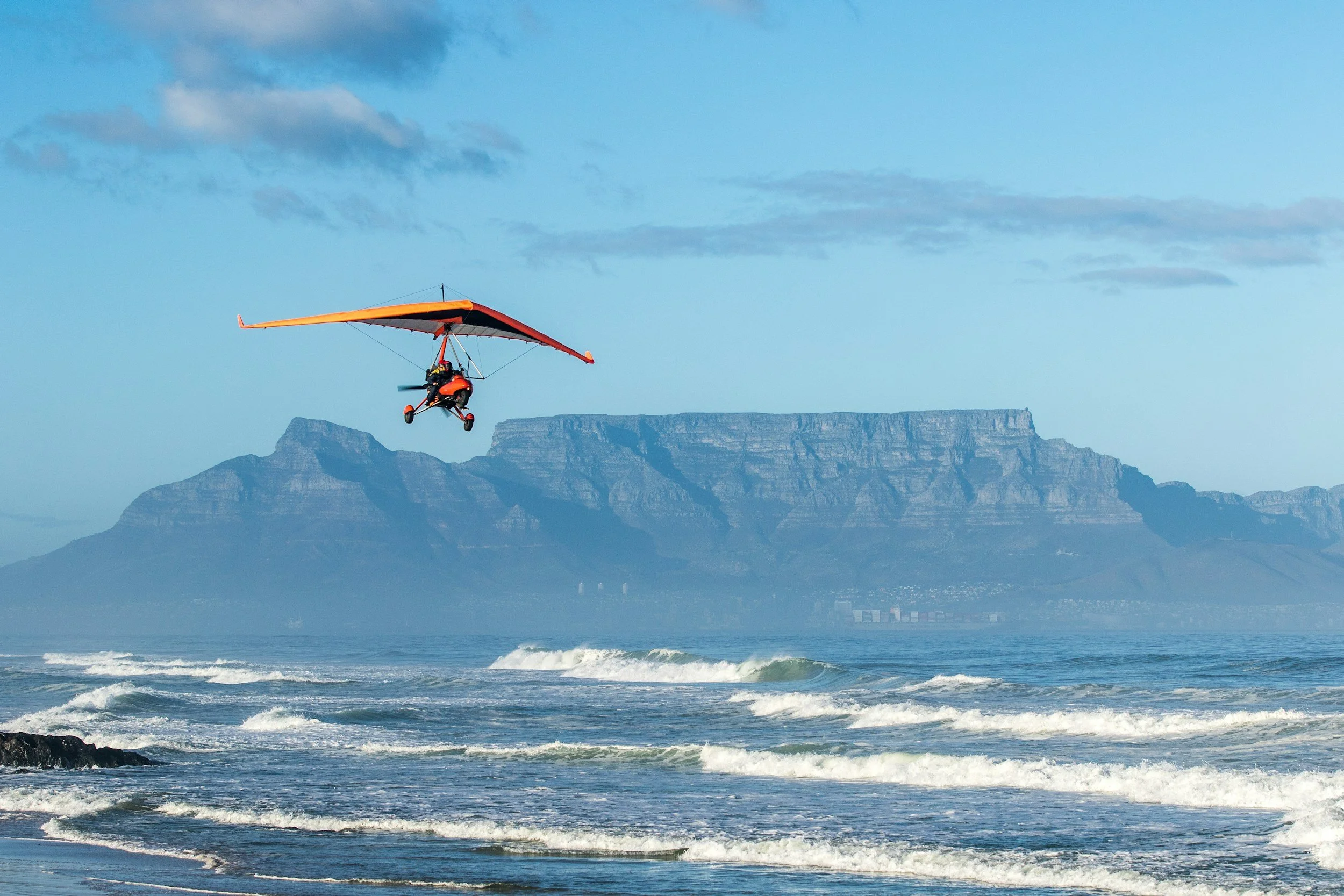 A person flying a powered hang glider over the ocean with waves and a mountain range in the background.