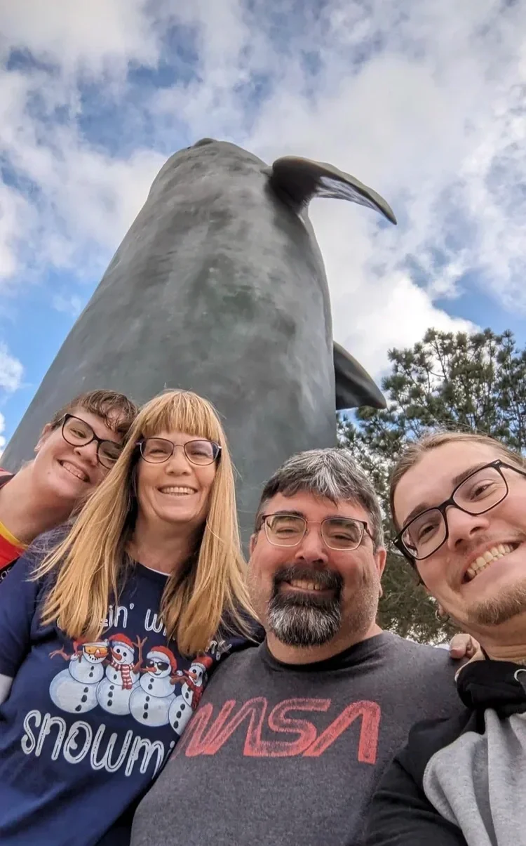 A group of four smiling people taking a selfie in front of a large sculpture of a whale's tail and body outdoors on a partly cloudy day.