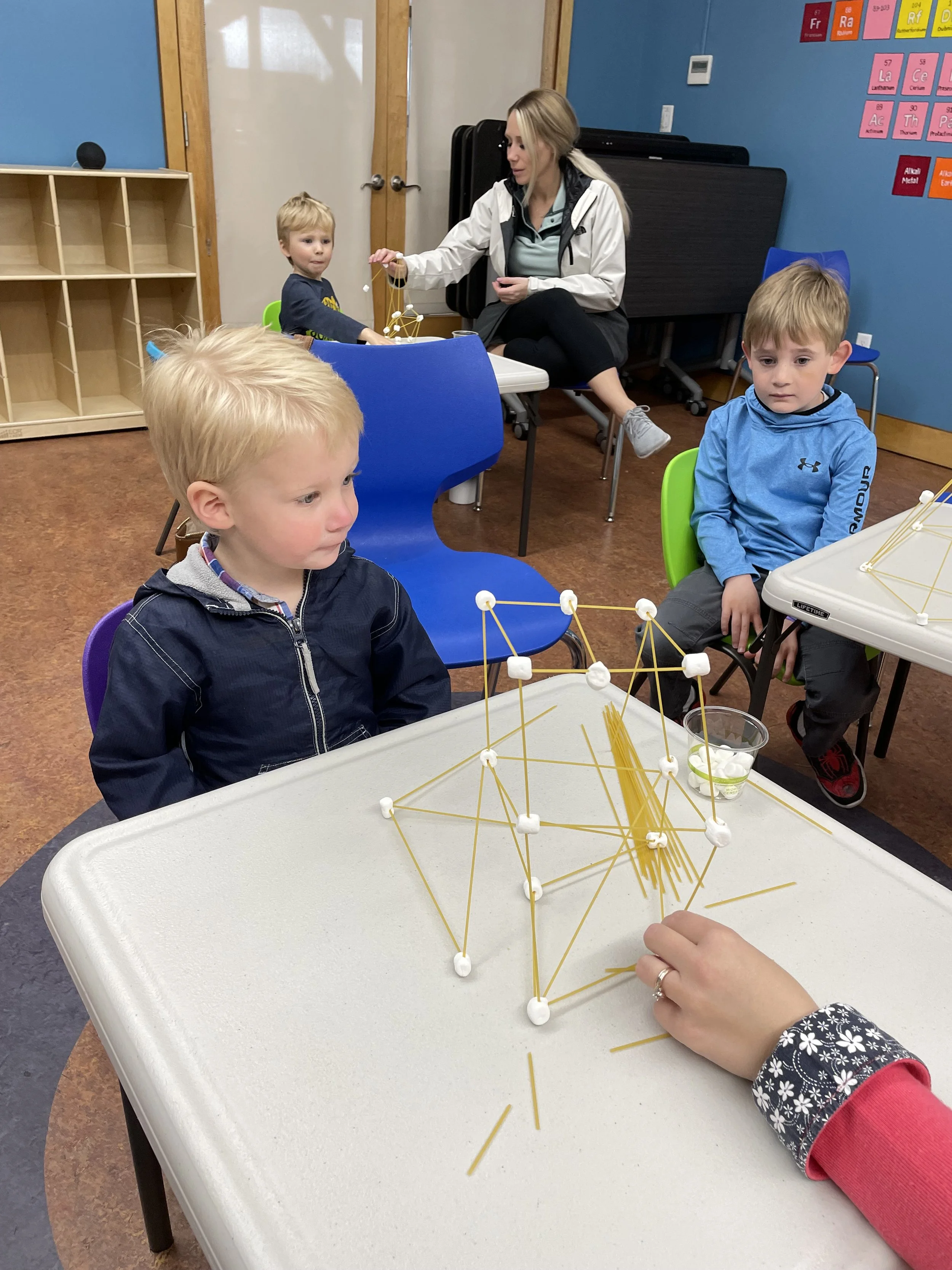 Children in a classroom building structures with spaghetti and marshmallows, supervised by an adult.